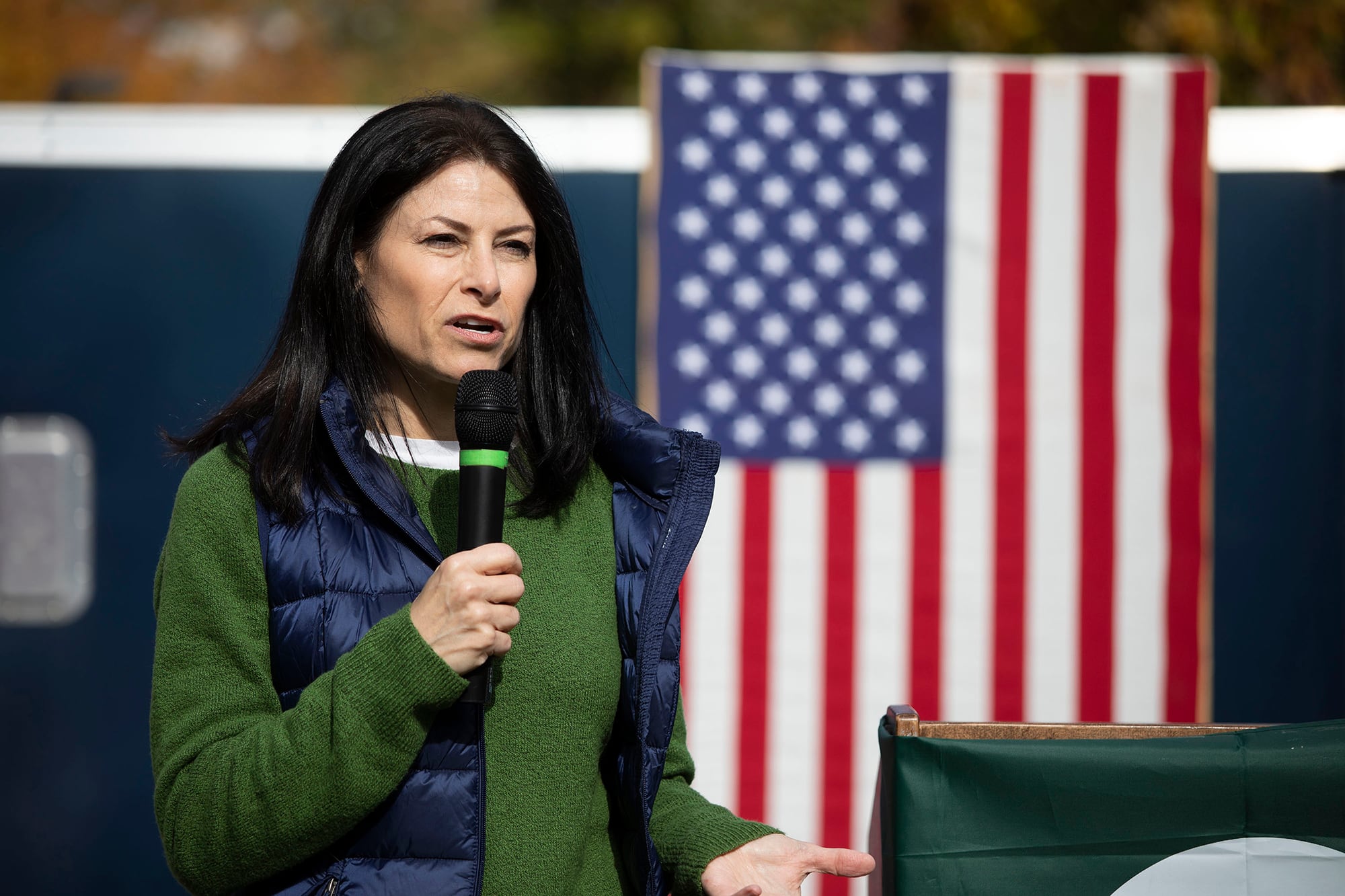 A white woman with dark hair and wearing a green shirt with a blue vest speaks in front of an American flag outside while holding a microphone.