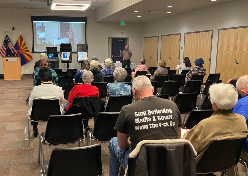 Two men stand in front of several rows of people sitting in chairs. There is a projector screen hanging from the wall in the background and a person wearing a shirt that reads "Sop believing media & govnt. Wake the F-ck Up" in the foreground.
