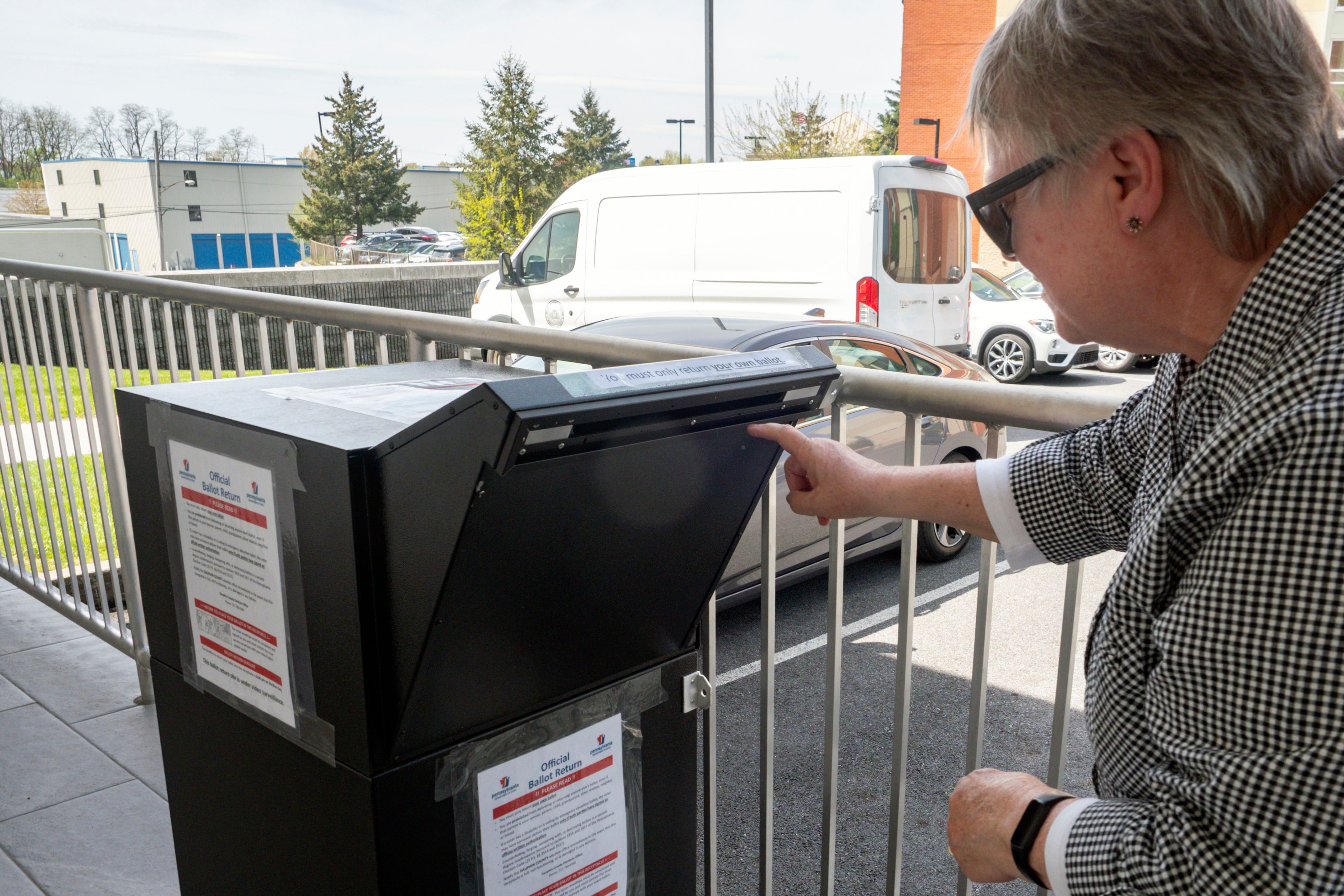 A person places a ballot into a box with a car in the background.