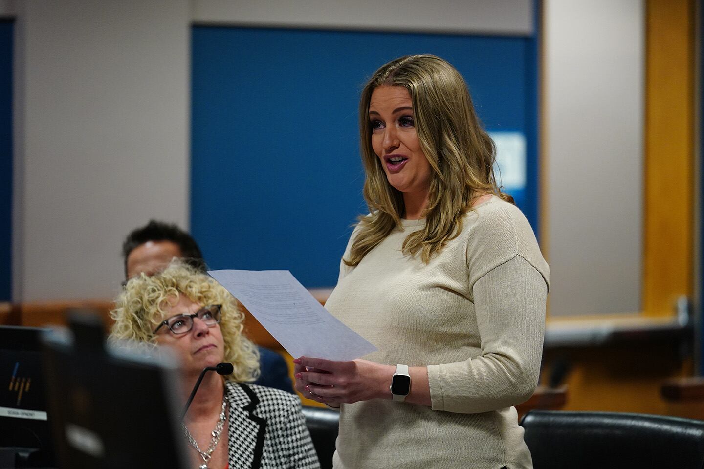 A women, standing up and wearing a white shirt, is reading from a piece of paper in a courtroom with a women sitting beside her.