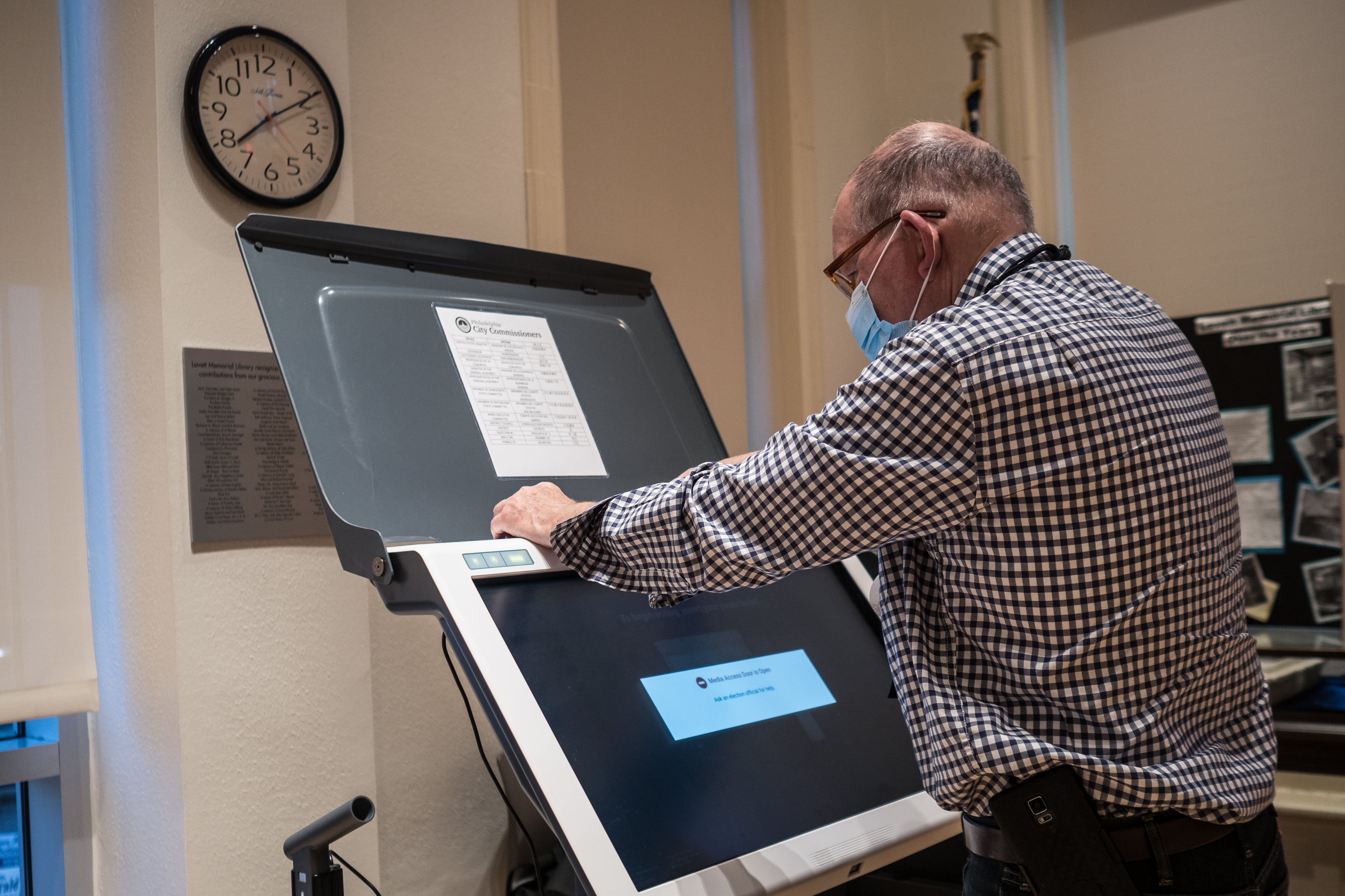 A photograph of a man wearing a mask and in a red dress shirt working at an ExpressVote machine inside of a room.