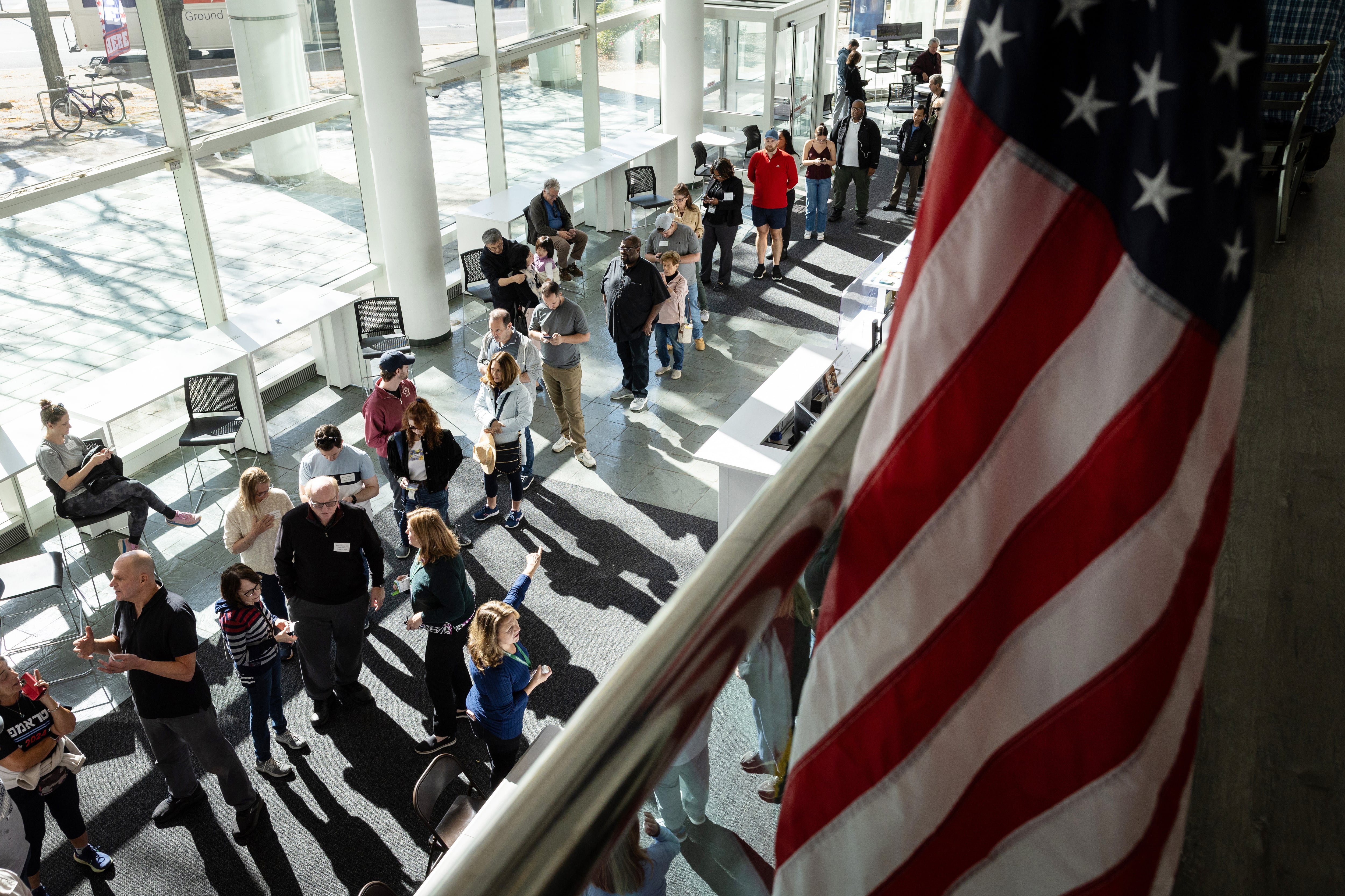 A long line of people stand in front of windows and an American flag sits off to the right above them.