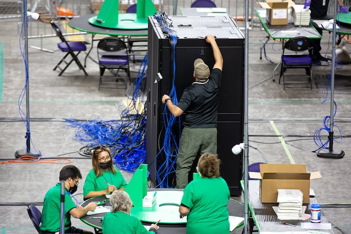 Four workers in green T-shirts seated at a round table on the floor of an arena. One man standing before a tall stack of computer servers reaches to the top of the equipment.