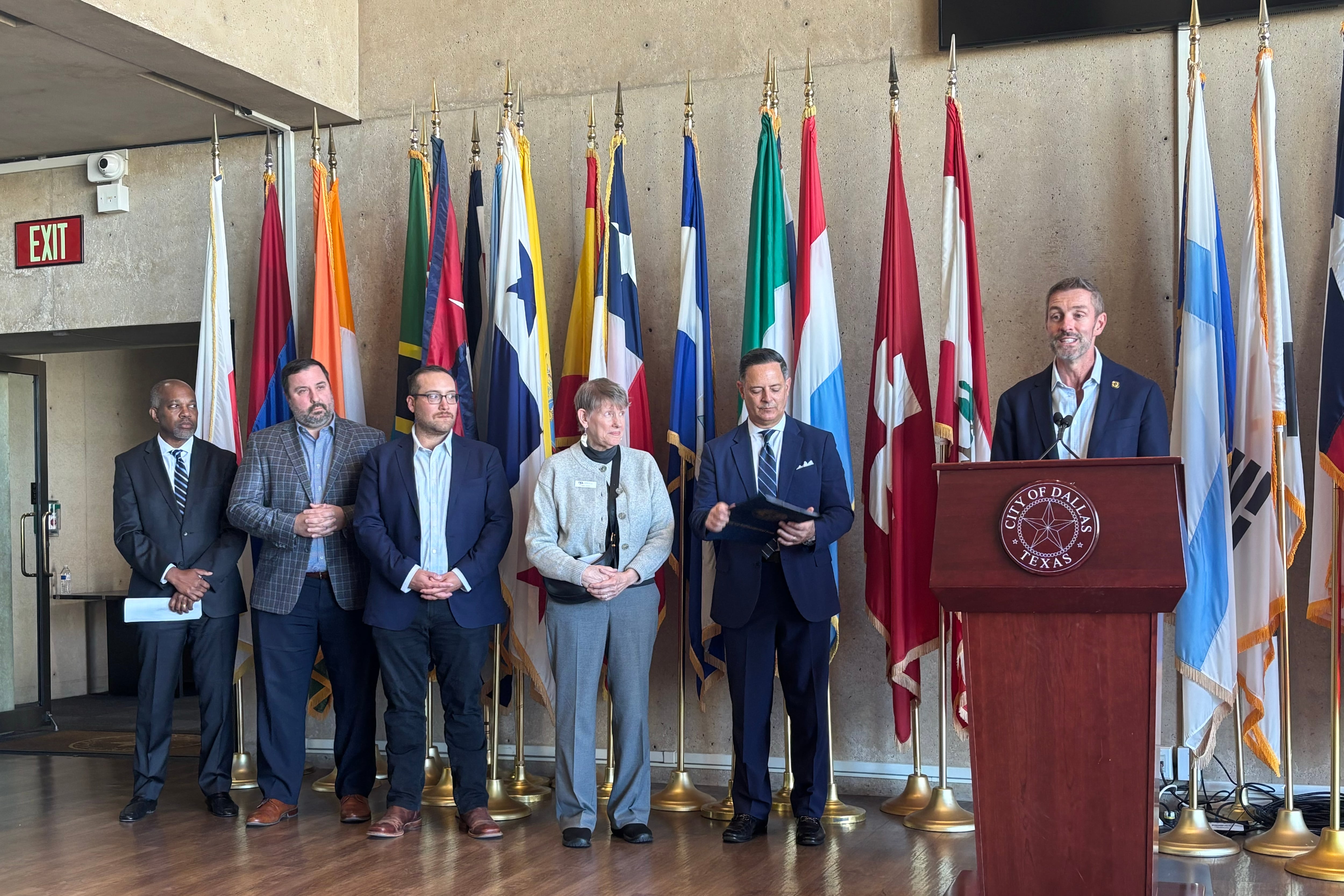 A photograph of a line of people in suits standing behind a podium and in front of a line of colorful flags.