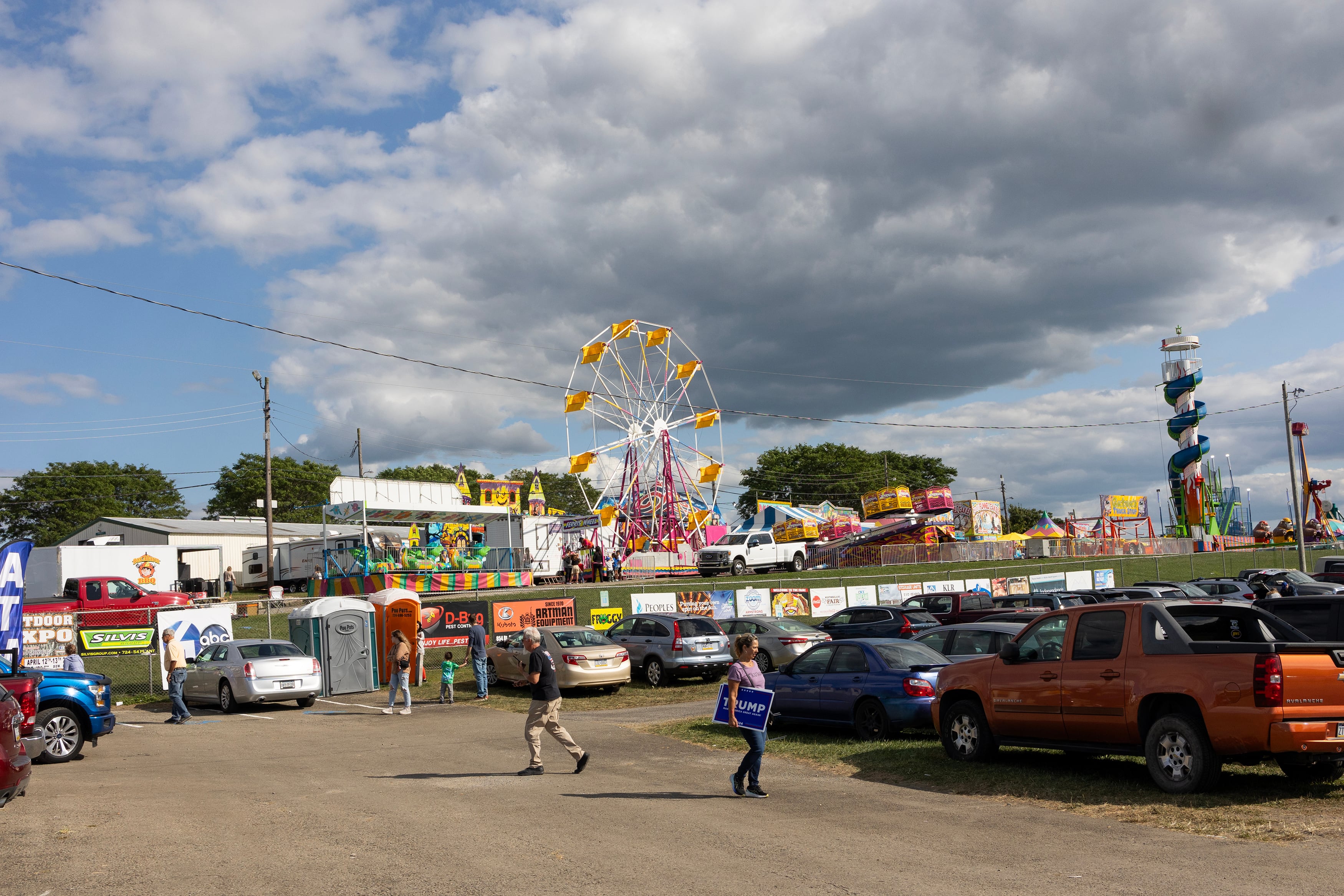 Two people walk across a dirt parking lot with a ferris wheel and other county fair tents and buildings in the background with a blue sky and large clouds in the sky.