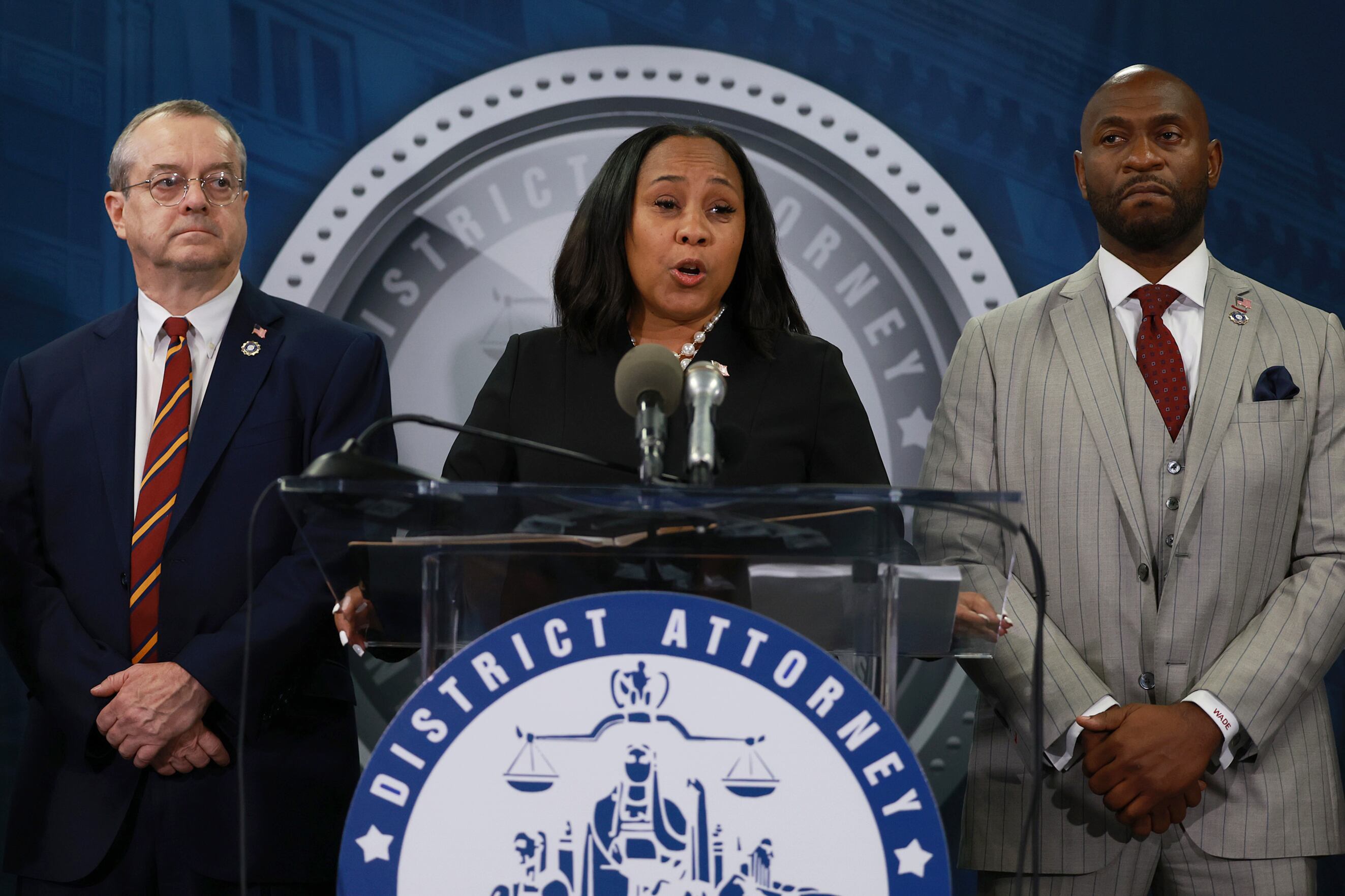 A woman in a dark suit speaks at a podium flanked by two men in suits.