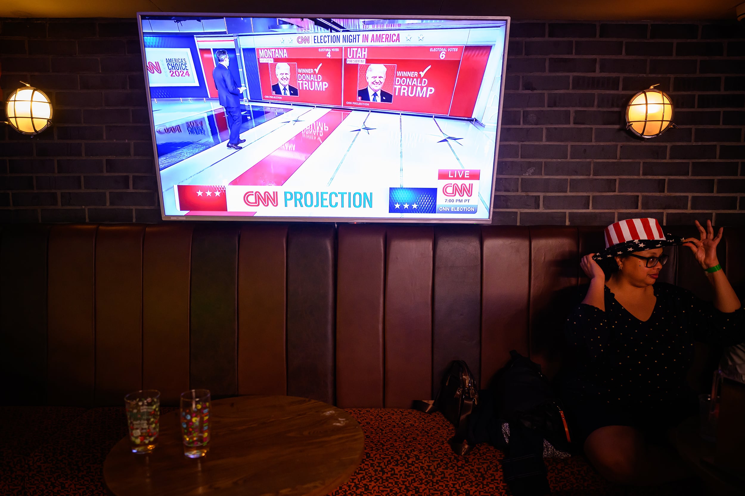 A photograph of a person sitting at a booth in a dark restaurant putting an American patriotic hat on while a large TV screen above her shows Donald Trump winning the 2024 presidential election.