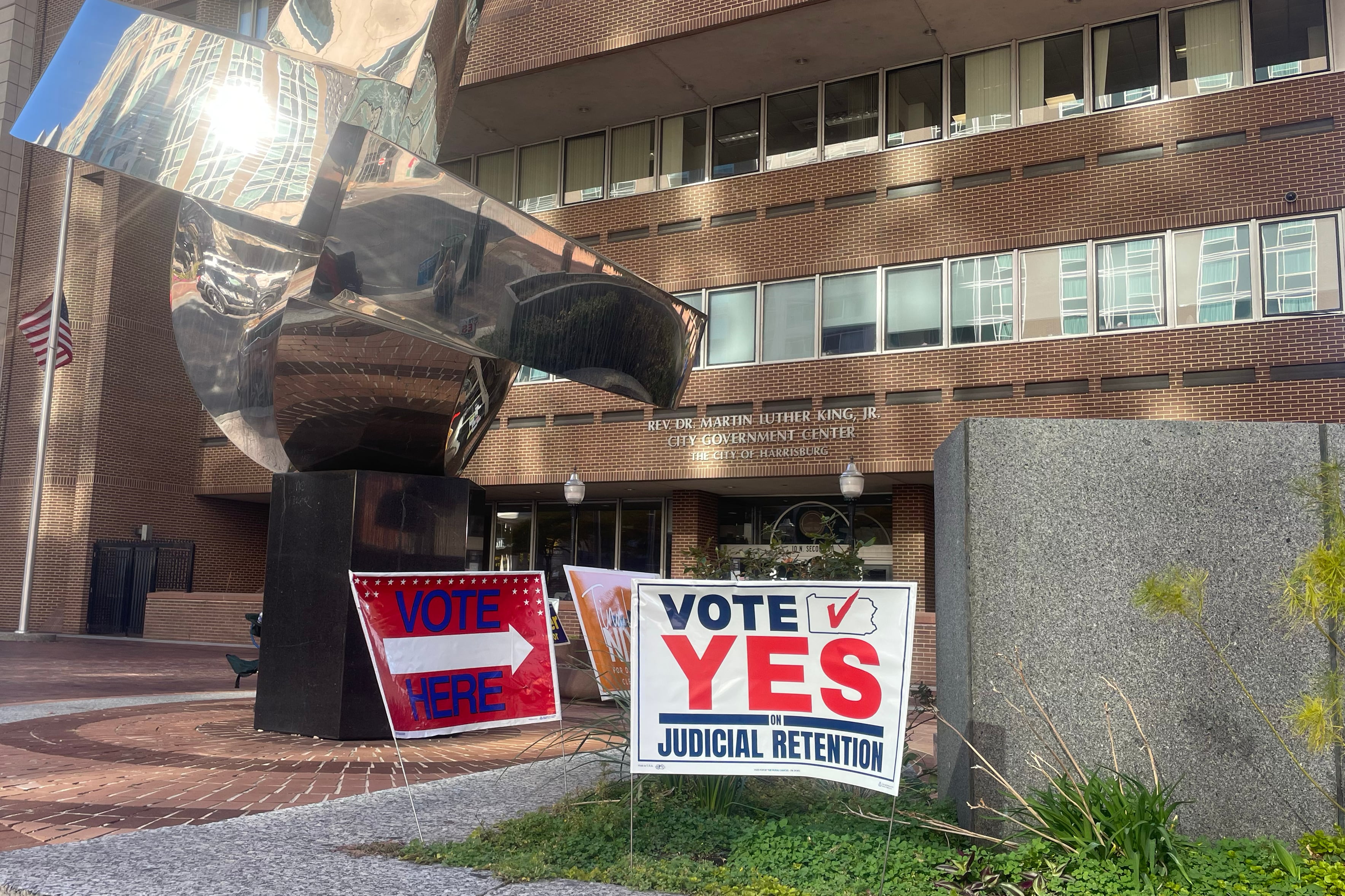 A photograph of voting signs on a small grassy area next to a large sculpture outside of a large brick building.