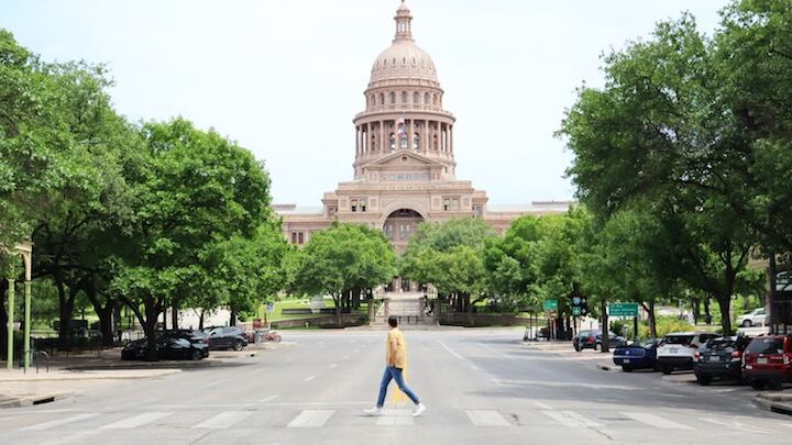 A man uses a crosswalk to cross a street and in the background is a statehouse building