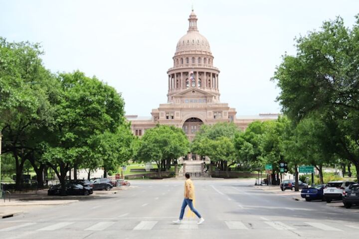 A man uses a crosswalk to cross a street and in the background is a statehouse building