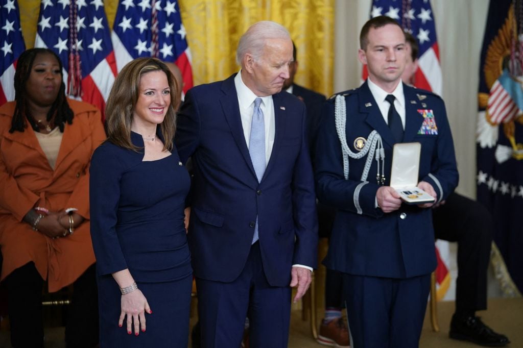 A man in a navy blue suit stands with his arm around a blonde woman in a blue dress, with a woman in an orange suit jacket standing against a backdrop of American flags and a uniformed man in the background. 