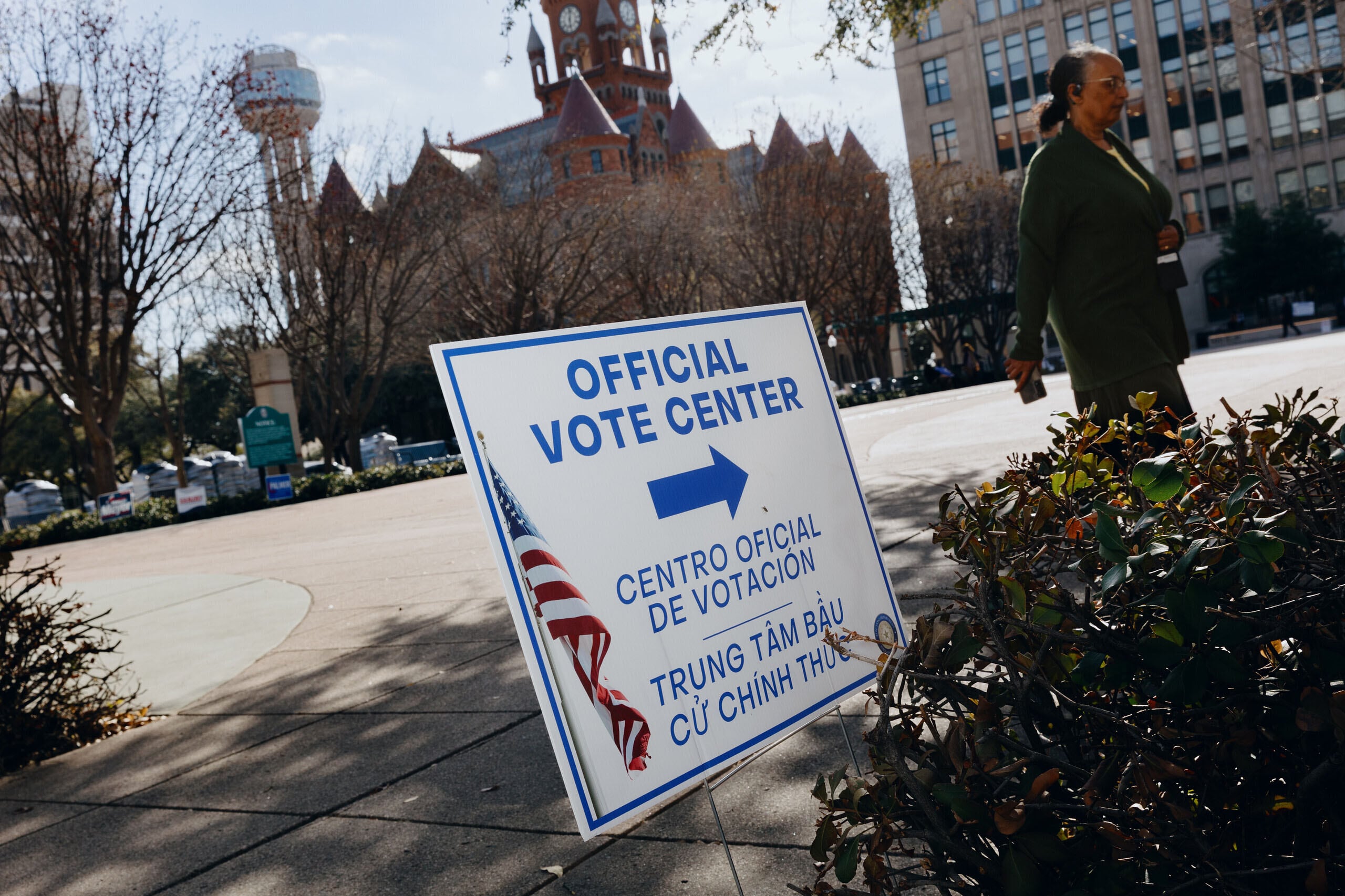 A photograph of a person walking by a sign that reads "Official Vote Center," outside on a sunny day.