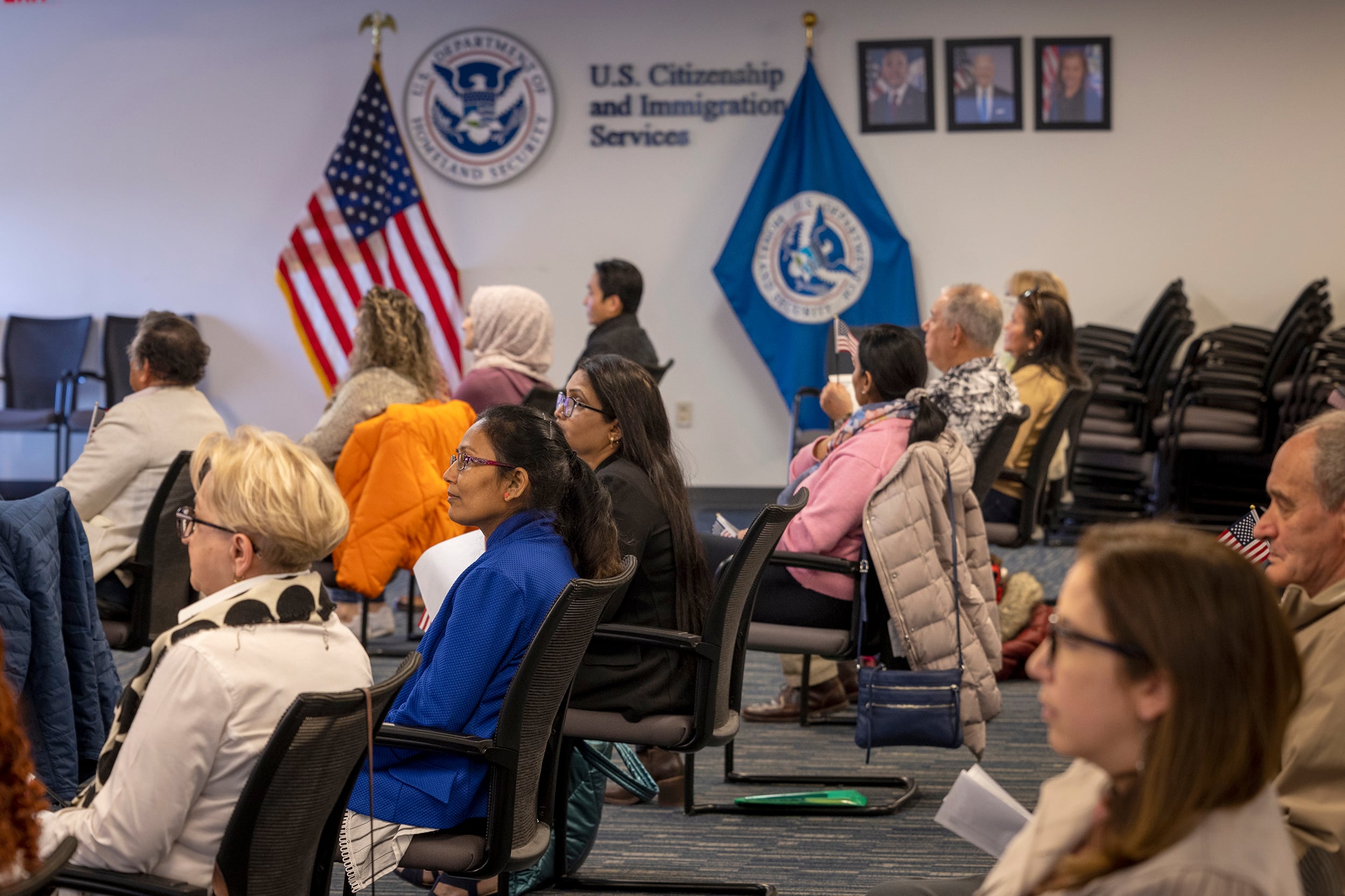 A photograph of a group of people sitting in chairs in a meeting room looking all in the same direction. In the background, there's an American flag and a USCIS flag.
