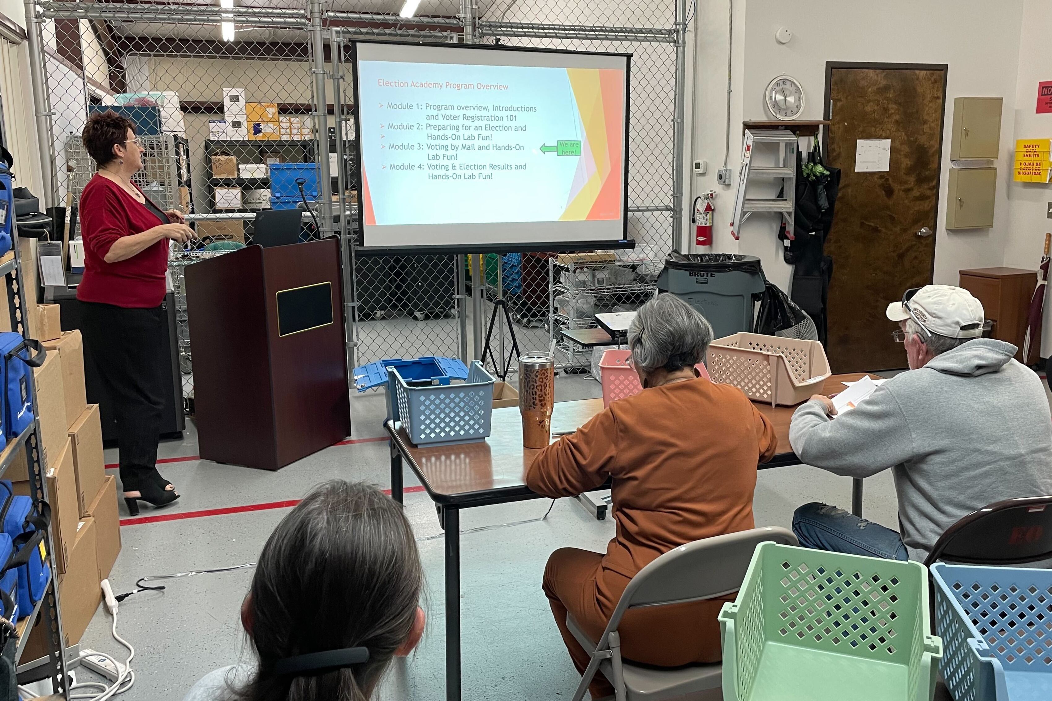 a group of people sit at desks looking at one woman who is giving a presentation at the front of the room next to a projector screen.