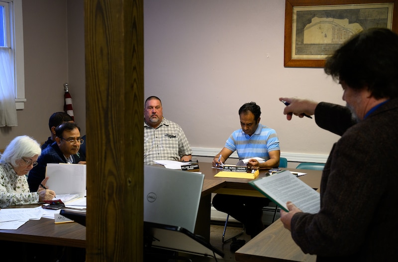 Four people sit along a table in a conference room while a man on the far right of the frame points to the group of people while reading from a piece of paper.