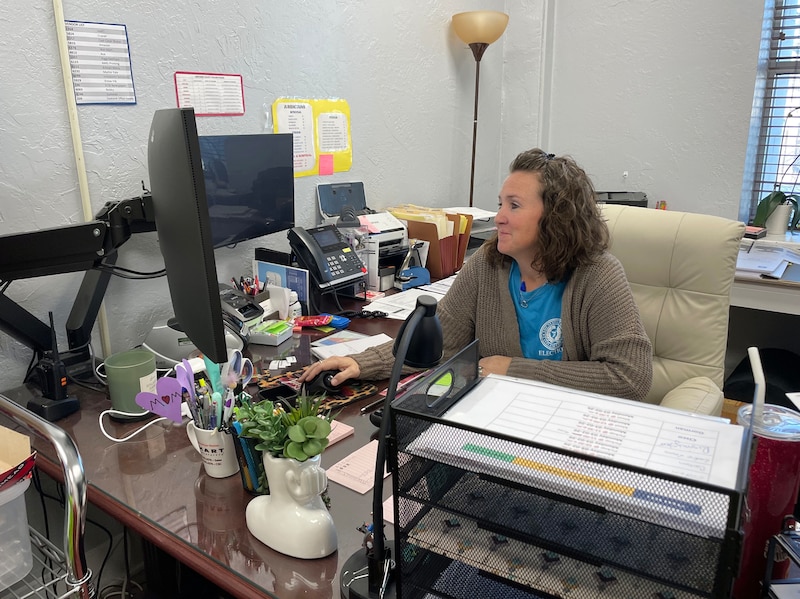 A photograph of a woman at a desk full of papers, books, a computer and some decorations.