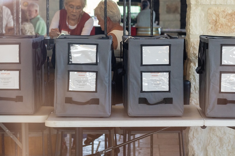 A row of ballot boxes is seen through a window with two people in the background.