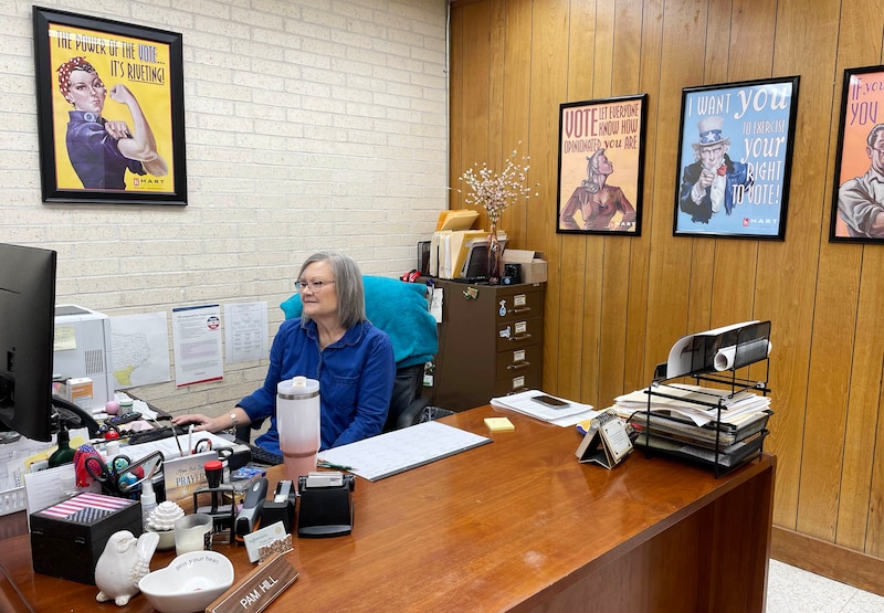A woman with short light hair and wearing a blue blouse sits behind a wooden desk with large posters on the wall in the background.