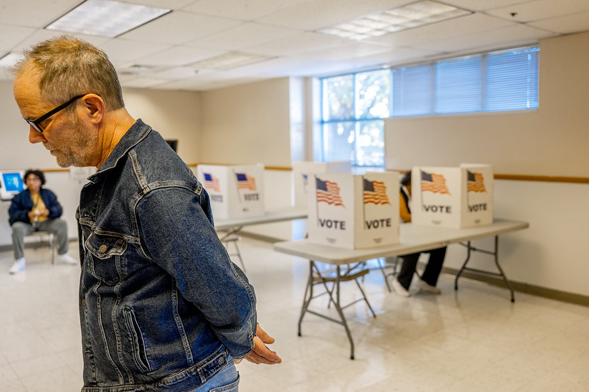 A man wearing a jean jacket walks in front of tables with dividers for people to vote in the background.