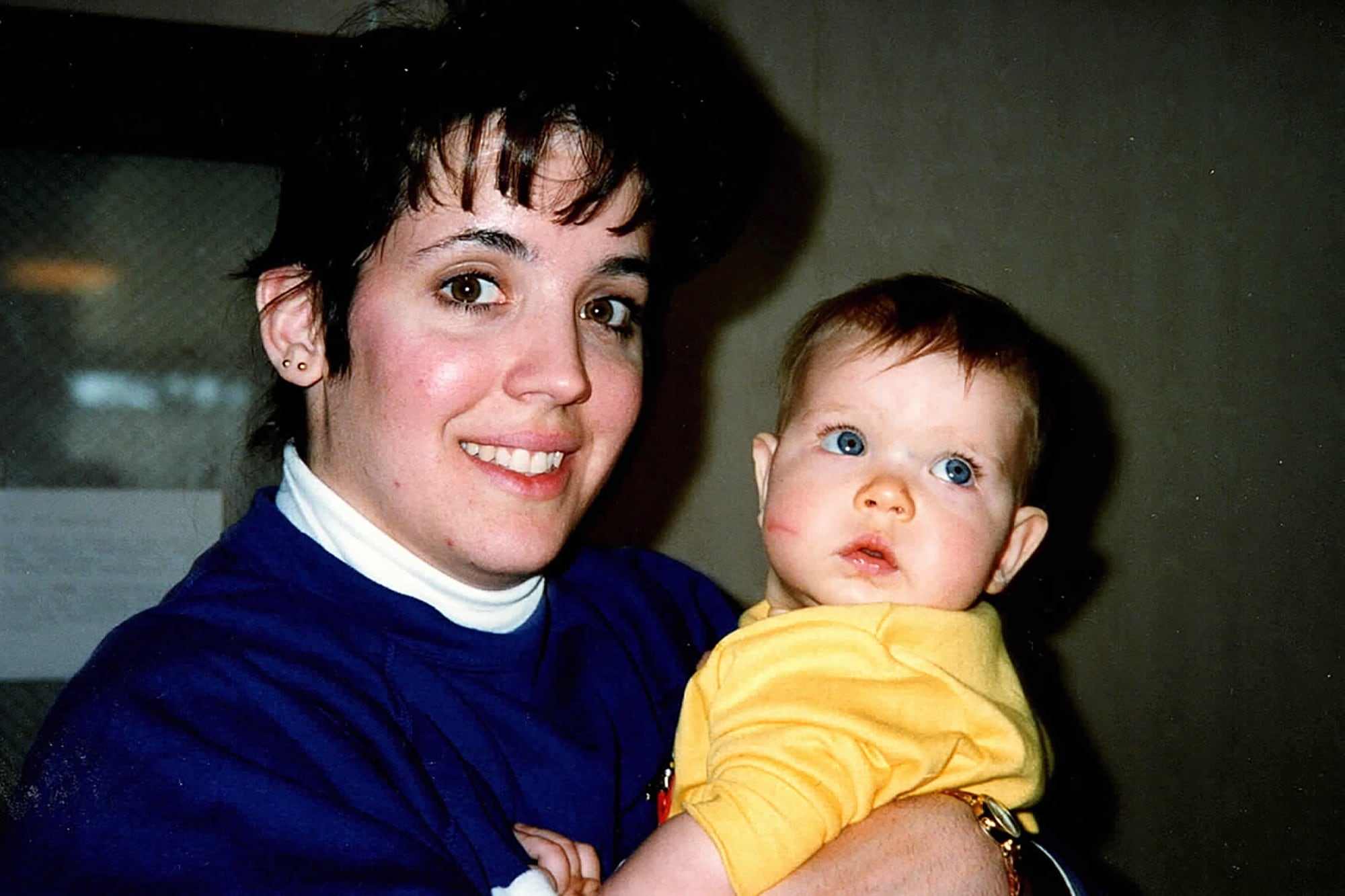 A woman with dark hair and wearing a blue sweater holds her baby daughter wearing a yellow shirt.
