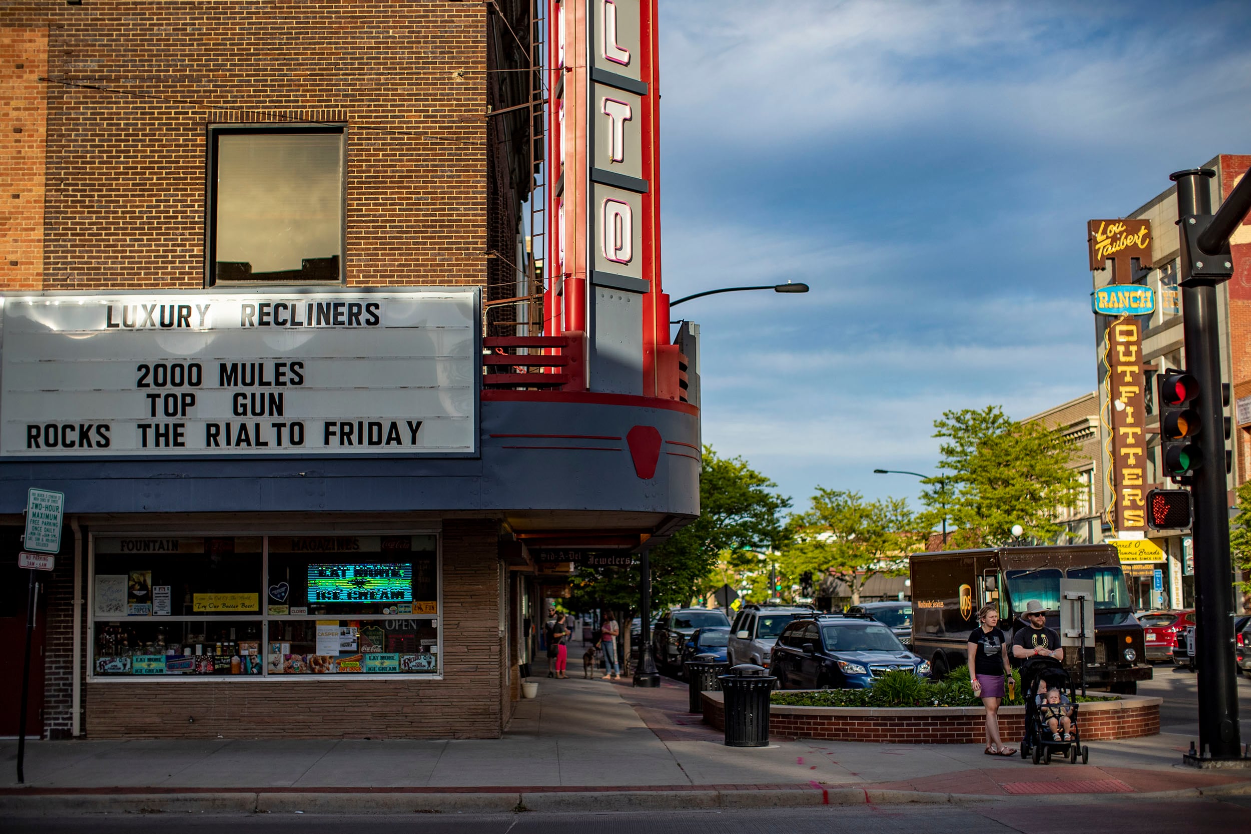Two people pushing a stroller stand at a crosswalk waiting to cross the street next to a movie theater marquee that has three movie titles including 2000 Mules and Top Gun.