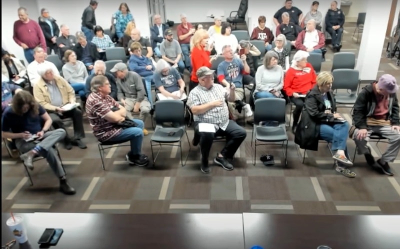 A group of people sit and stand in a couple of rows of chairs in a meeting room.