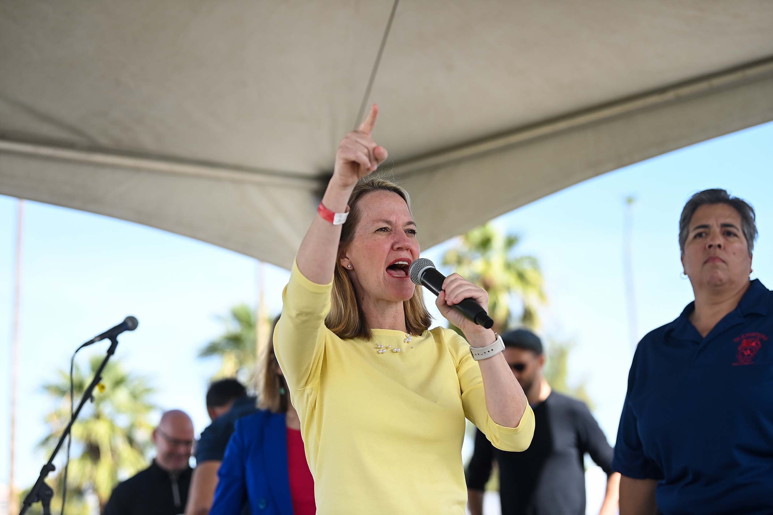 A photograph of a white woman in a green shirt speaking into a microphone under a tent while a few people stand behind her in the background.