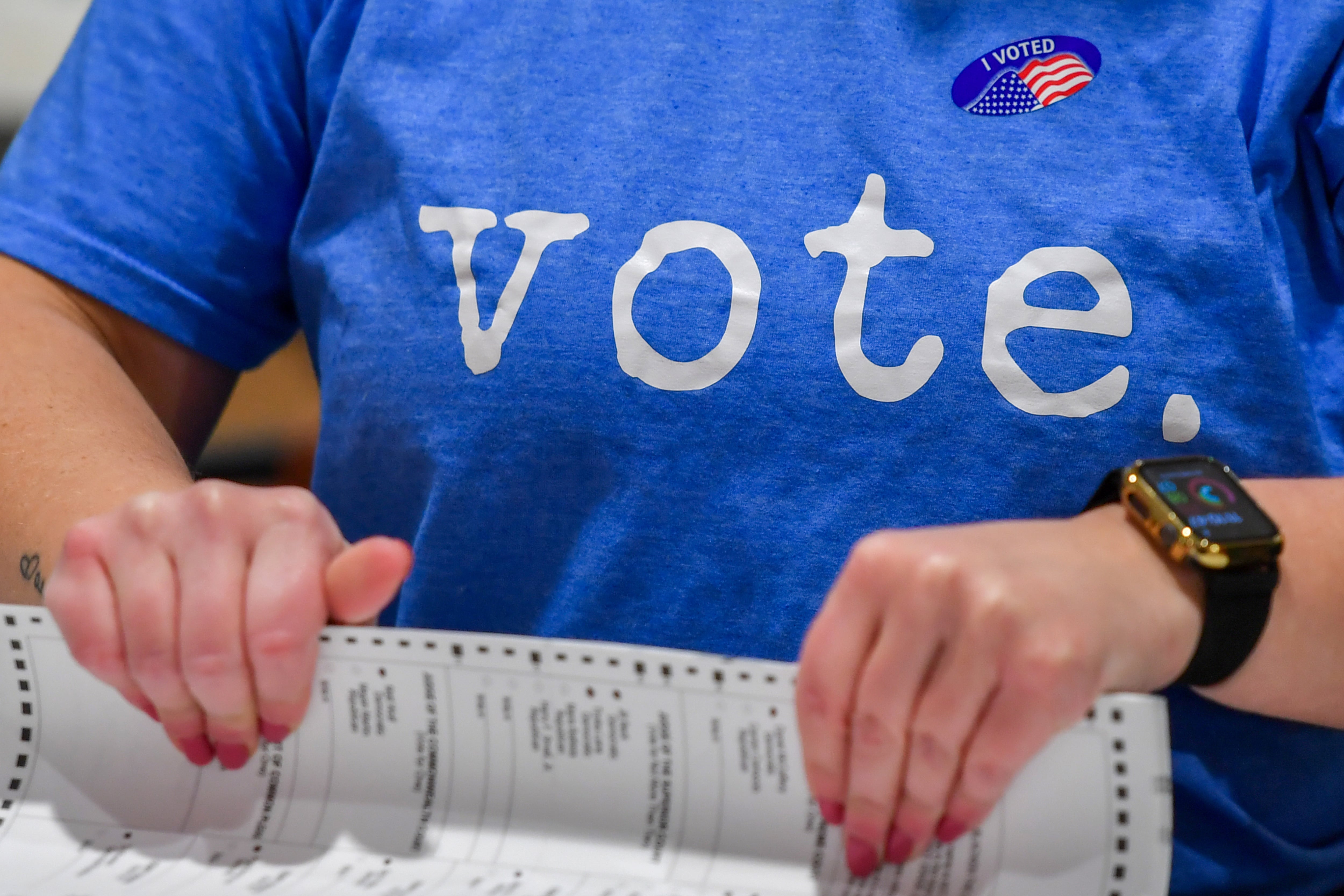 A closeup of a woman wearing a blue shirt that reads "vote" while holding a ballot