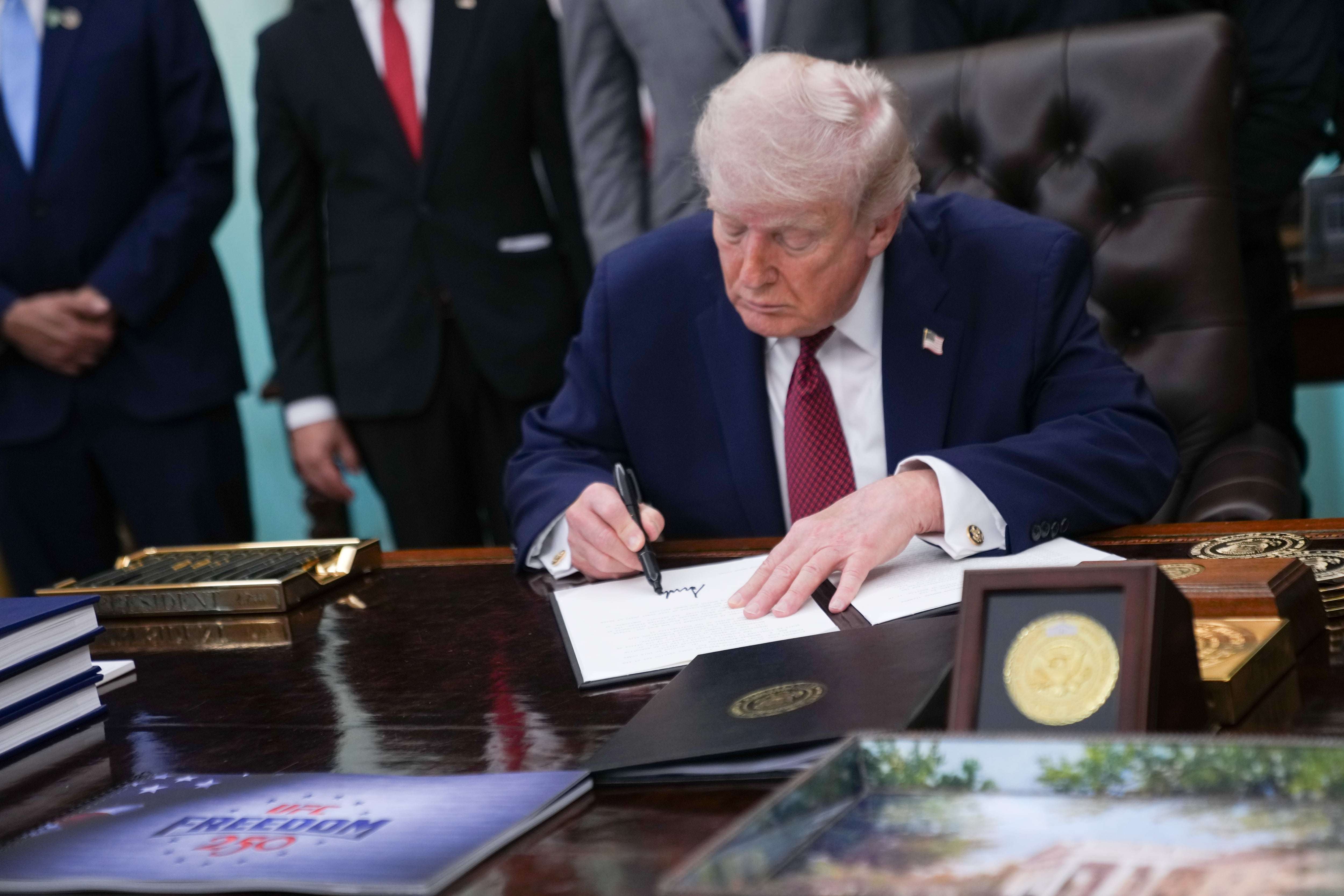 A photograph of President Donald Trump in a dark blue suit with a red tie sitting at a wooden table signing an executive order. There are people standing behind him but you can't see them.