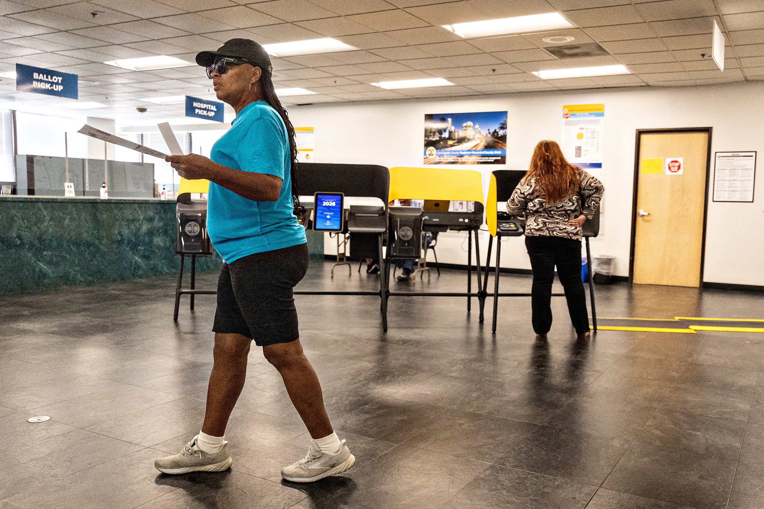 One person walks their ballot through a voting location while a person votes in the background.