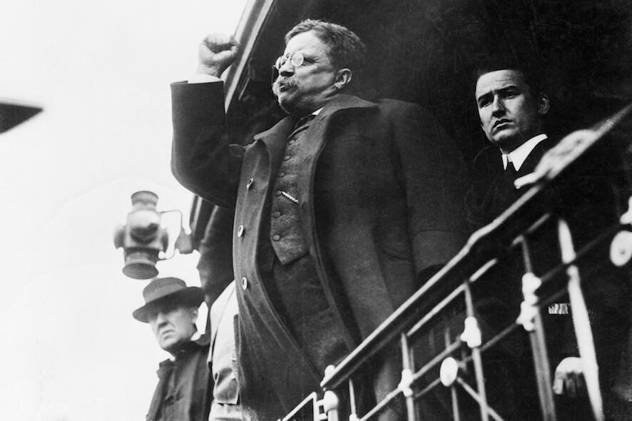 Black and white photograph of President Roosevelt standing at a railing with his fist raised aggressively.