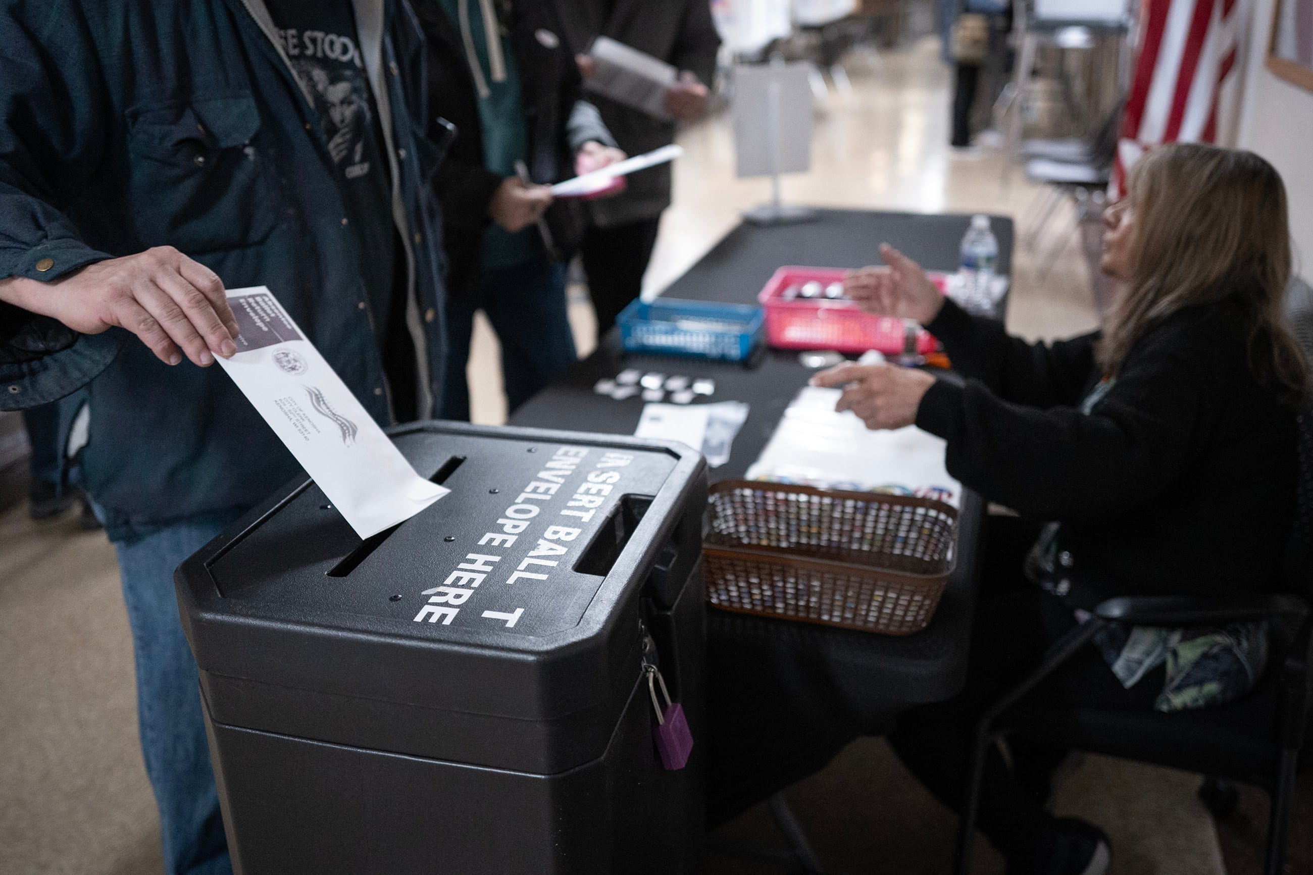 A person places an absentee ballot vote into a large black box that is sitting next to a table with a woman with long blonde hair sitting behind it. There is a line of people behind the person placing their ballot and a partial view of the hallway.