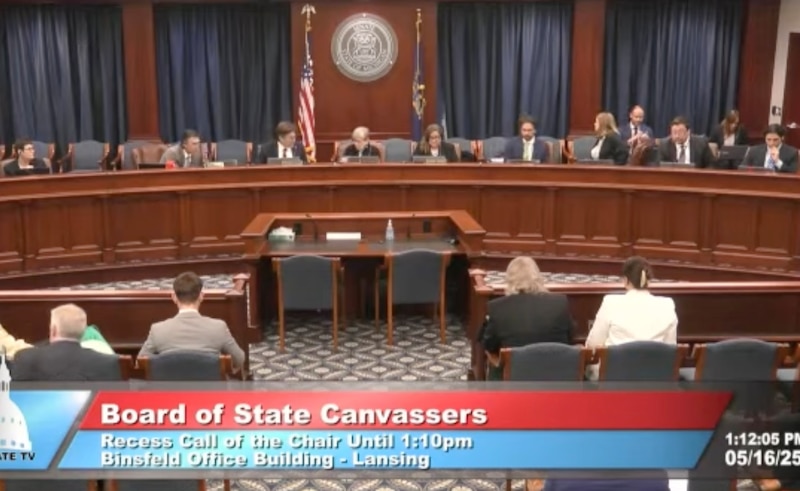 A screengrab showing a large group of people sitting behind a rounded wooden table in front of a row of people sitting in chairs.