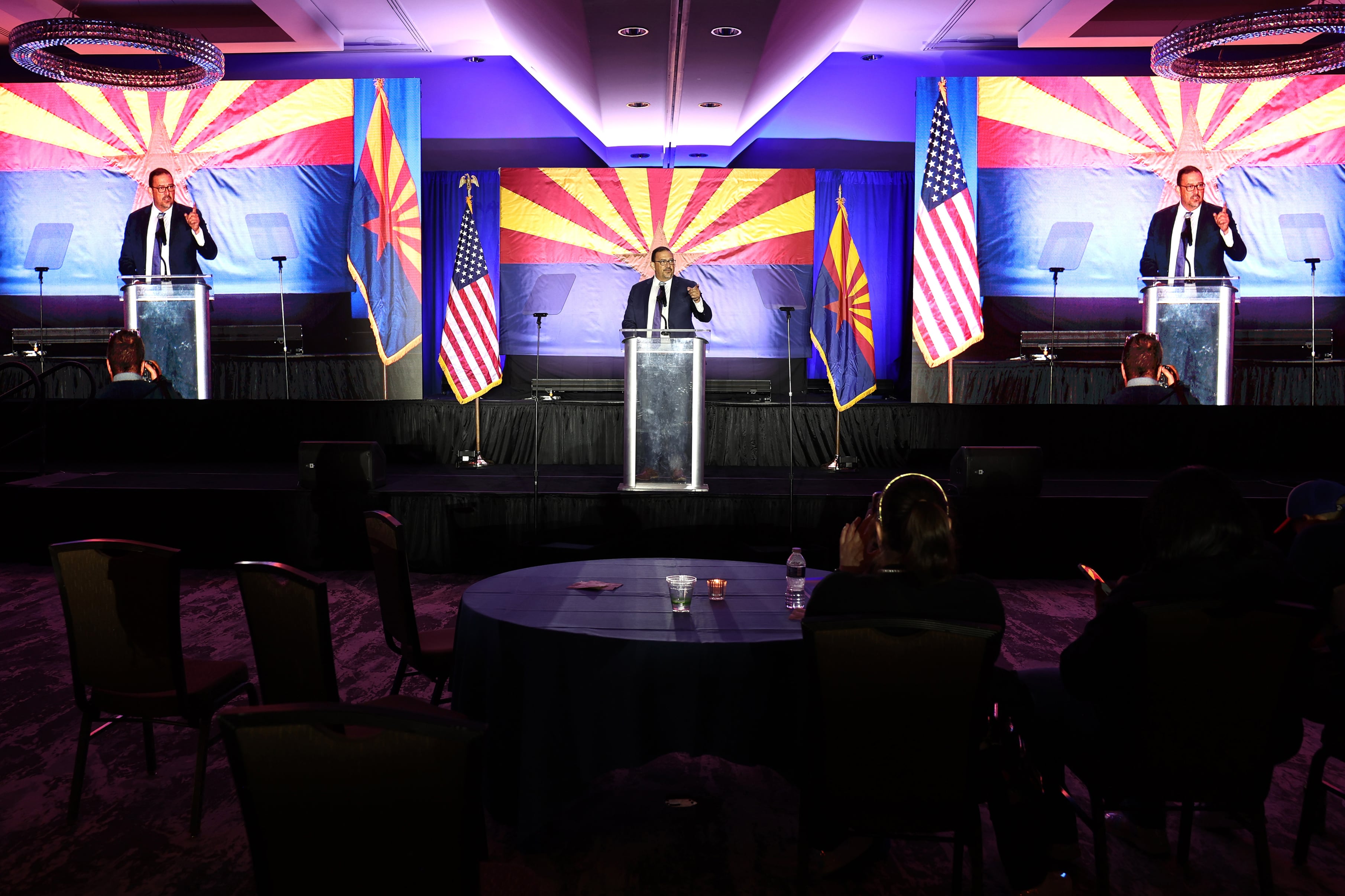 A man in a suit stands behind a podium and in between two large screens showing him speaking.