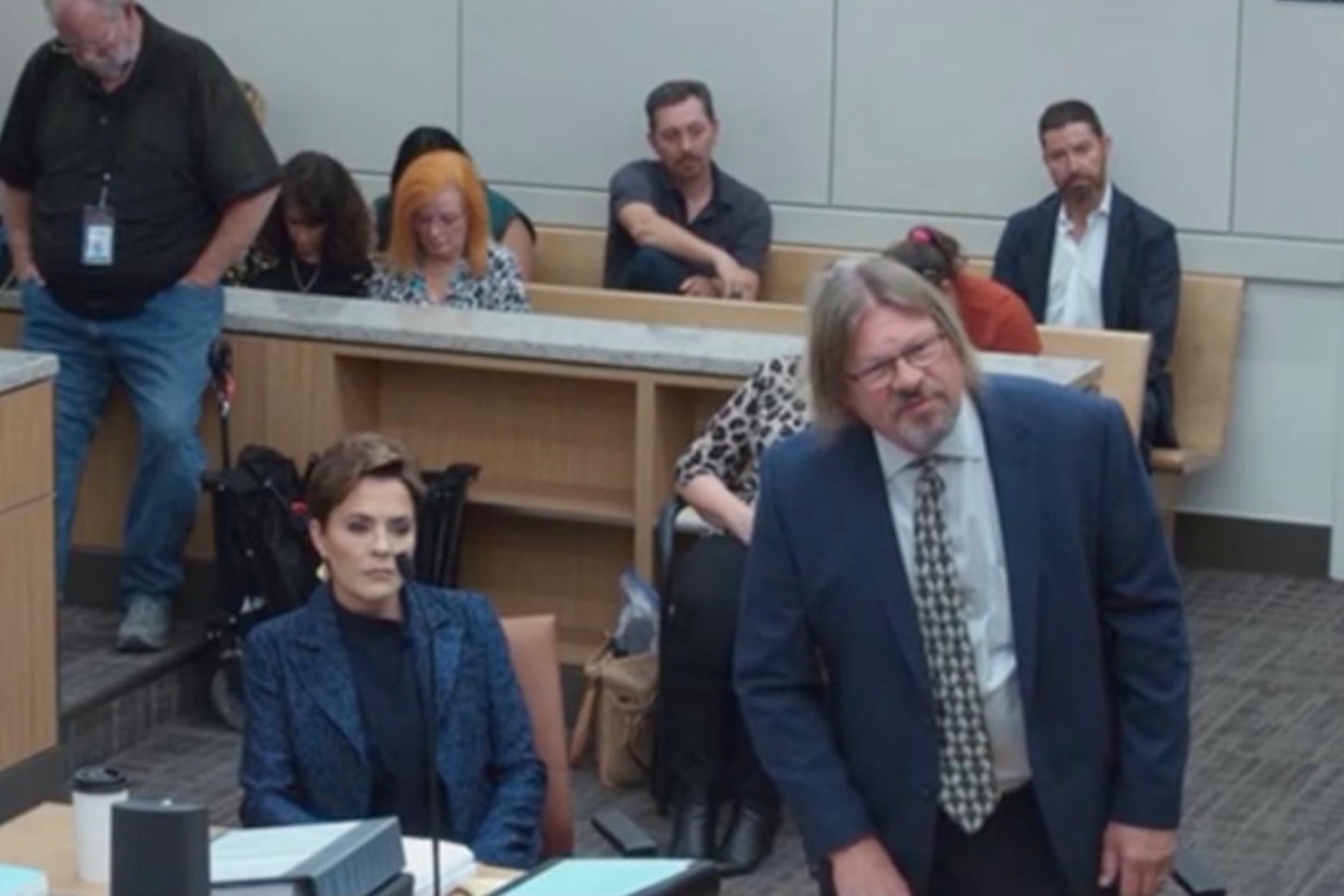 One woman sits while one man stands next to each other in a court room wearing business clothes with a group of people sitting in rows in the background.