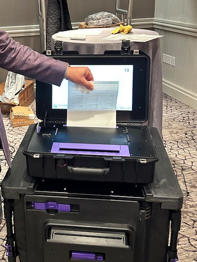 A hand holding a piece of paper over a voting machine in a conference room.