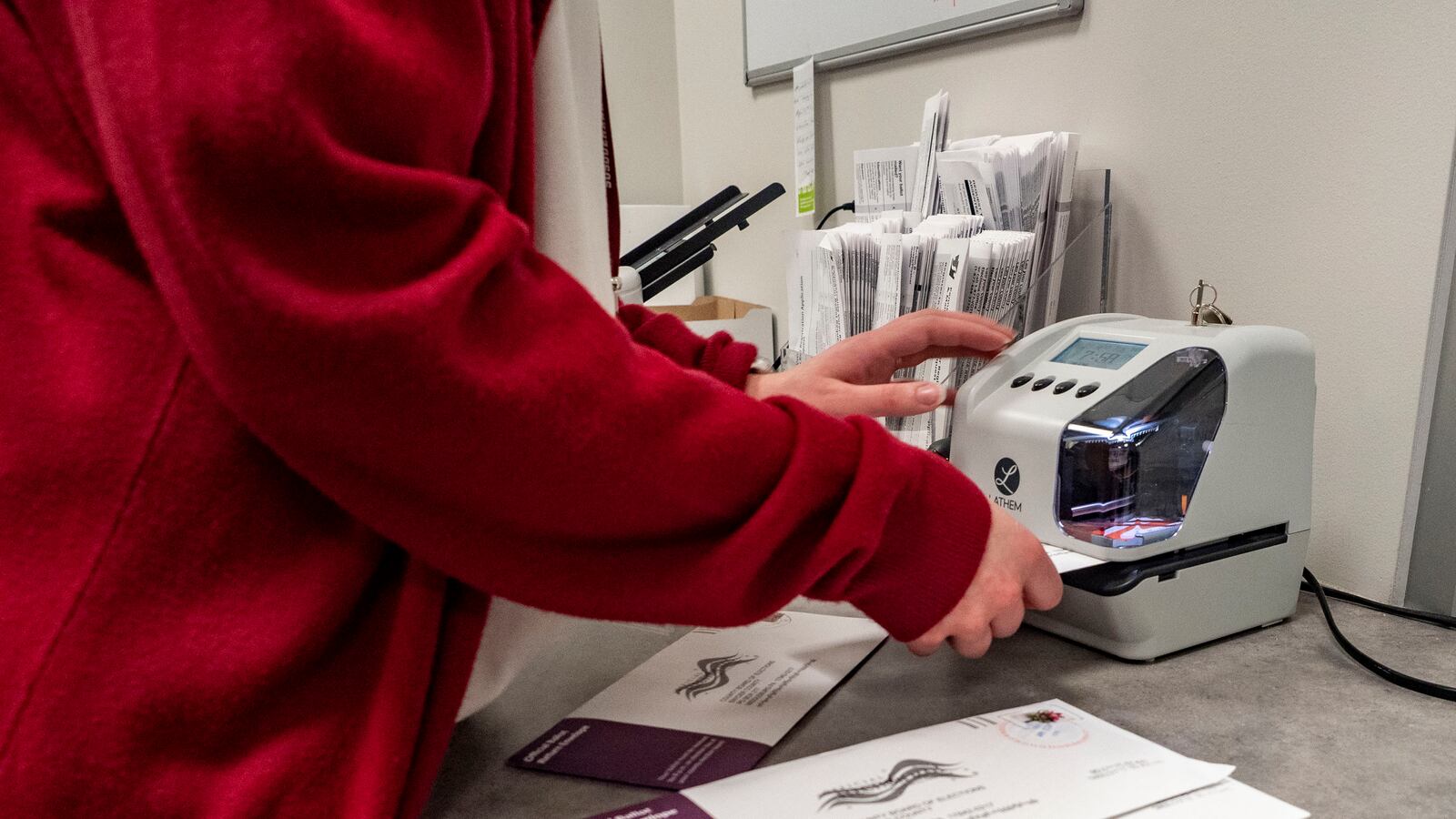 A person wearing a red sweater holds ballots and uses a small machine on a table.