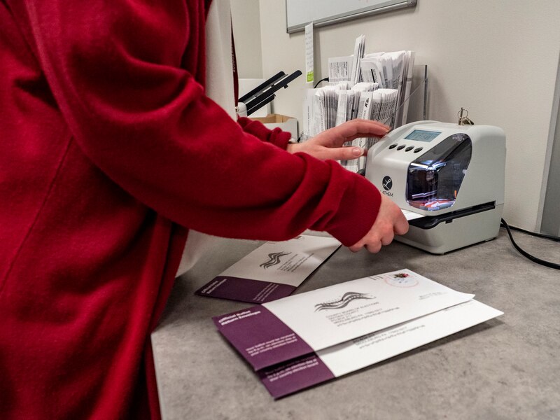 A person wearing a red sweater holds ballots and uses a small machine on a table.