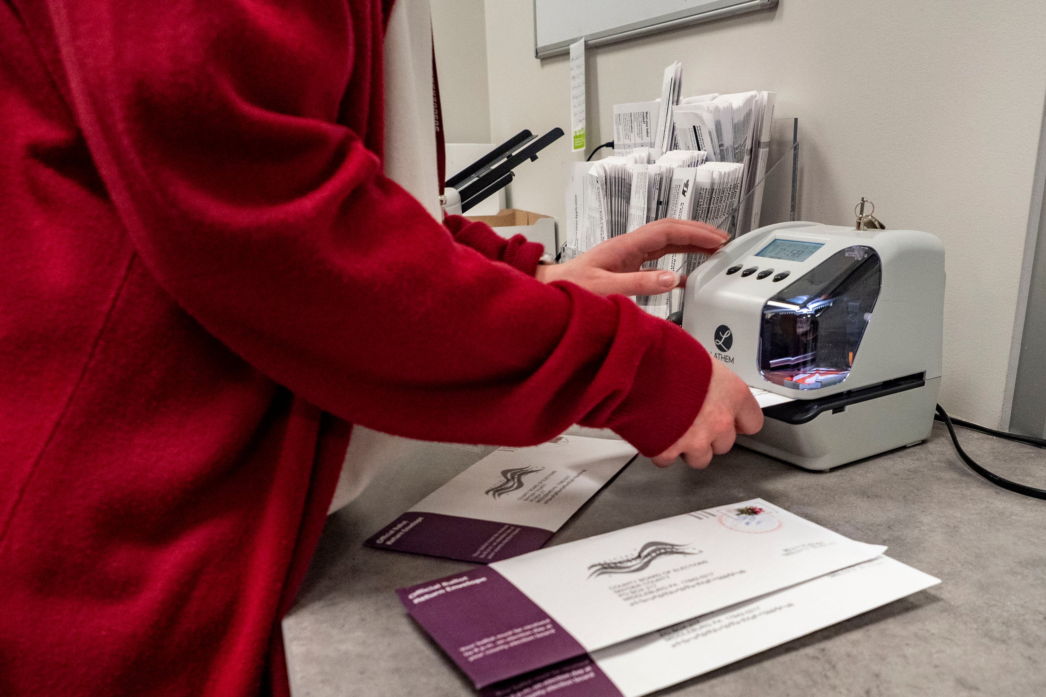 A person wearing a red sweater holds ballots and uses a small machine on a table.