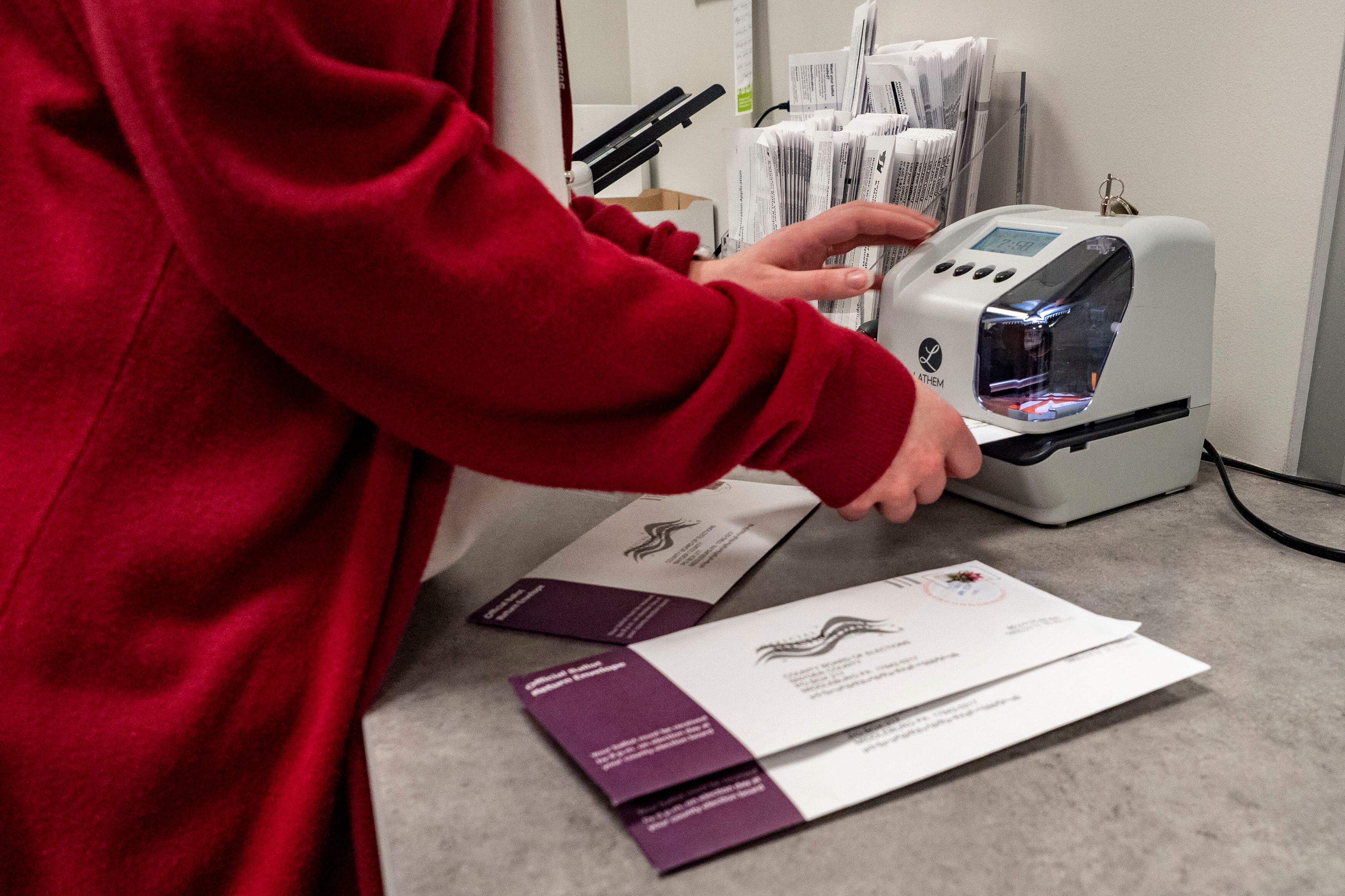 A person wearing a red sweater holds ballots and uses a small machine on a table.