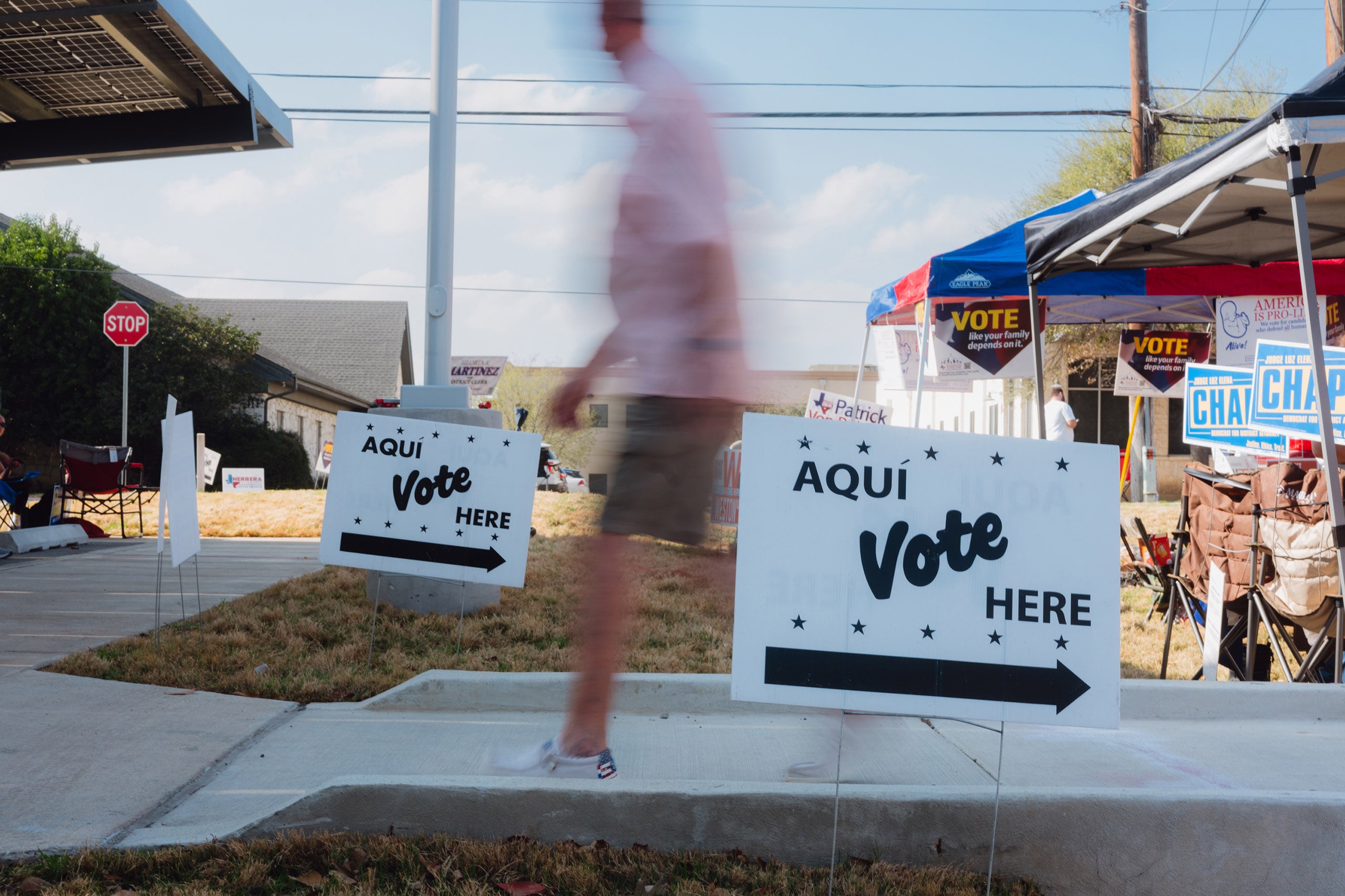 A person walks by two "vote here/aqui" signs outside on a sunny day.