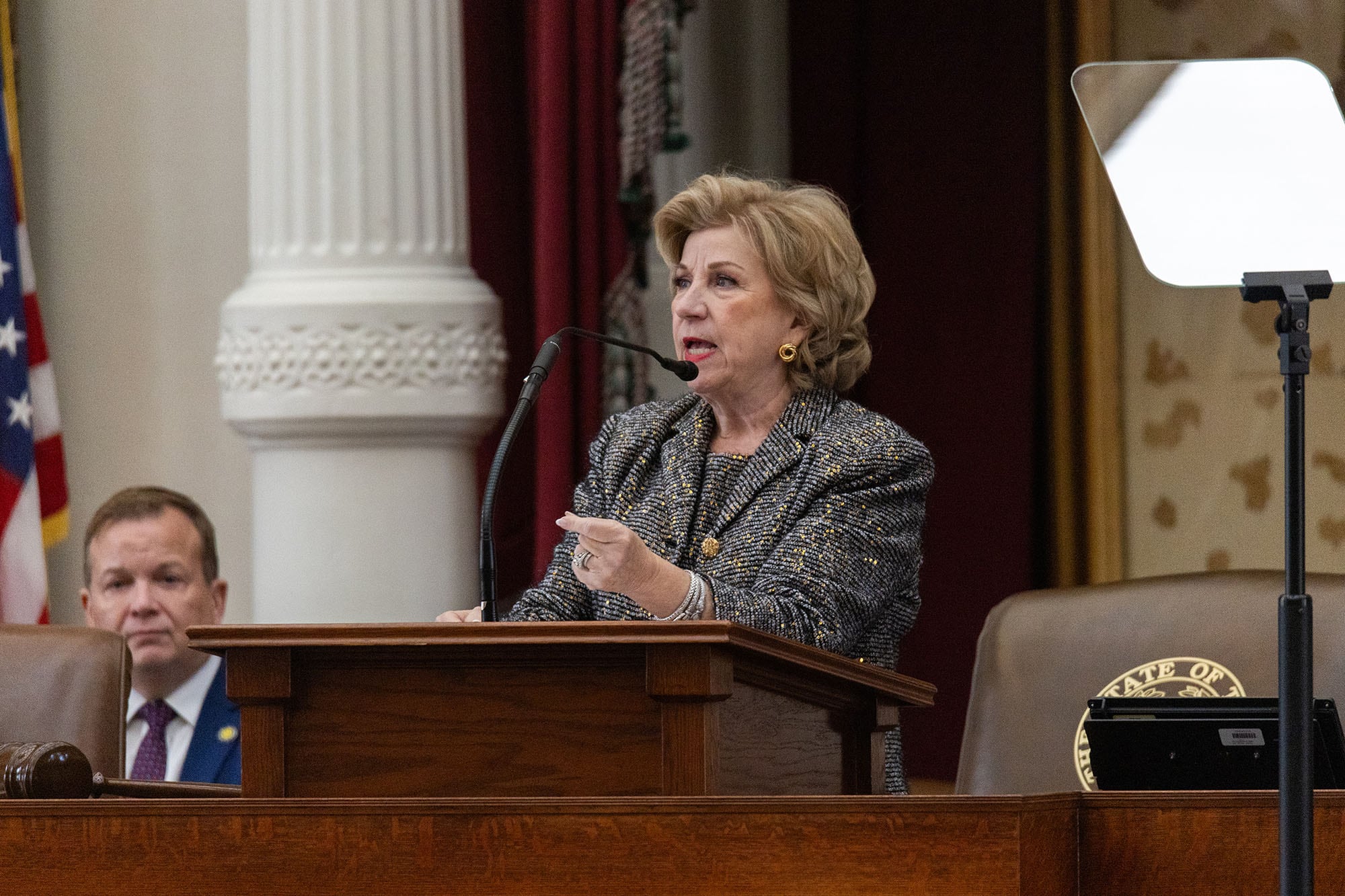 A photograph of a white woman speaking from a wooden podium.