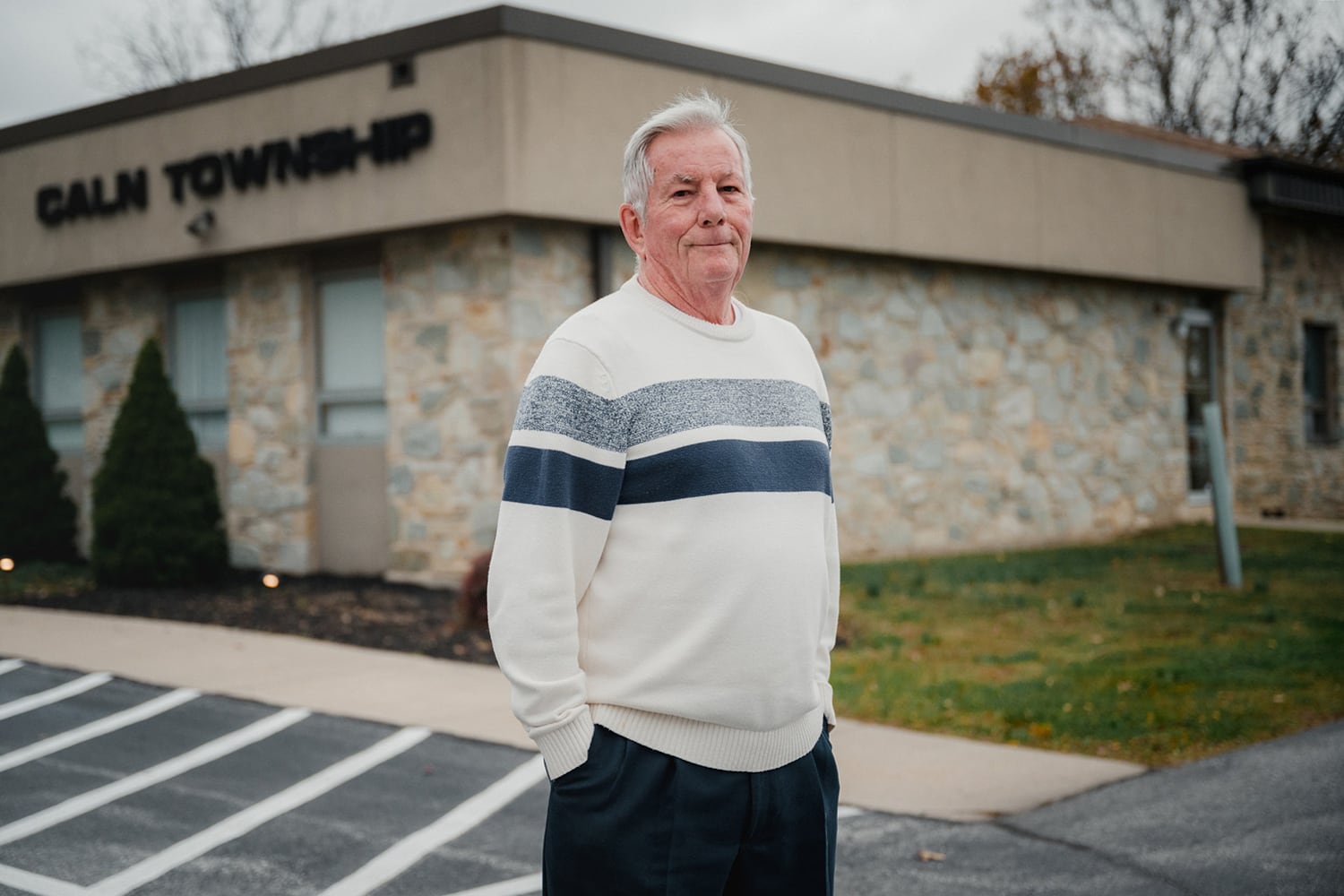 A man with white hair and wearing a white sweater with two blue stripes stands in front of a grey, brick building.