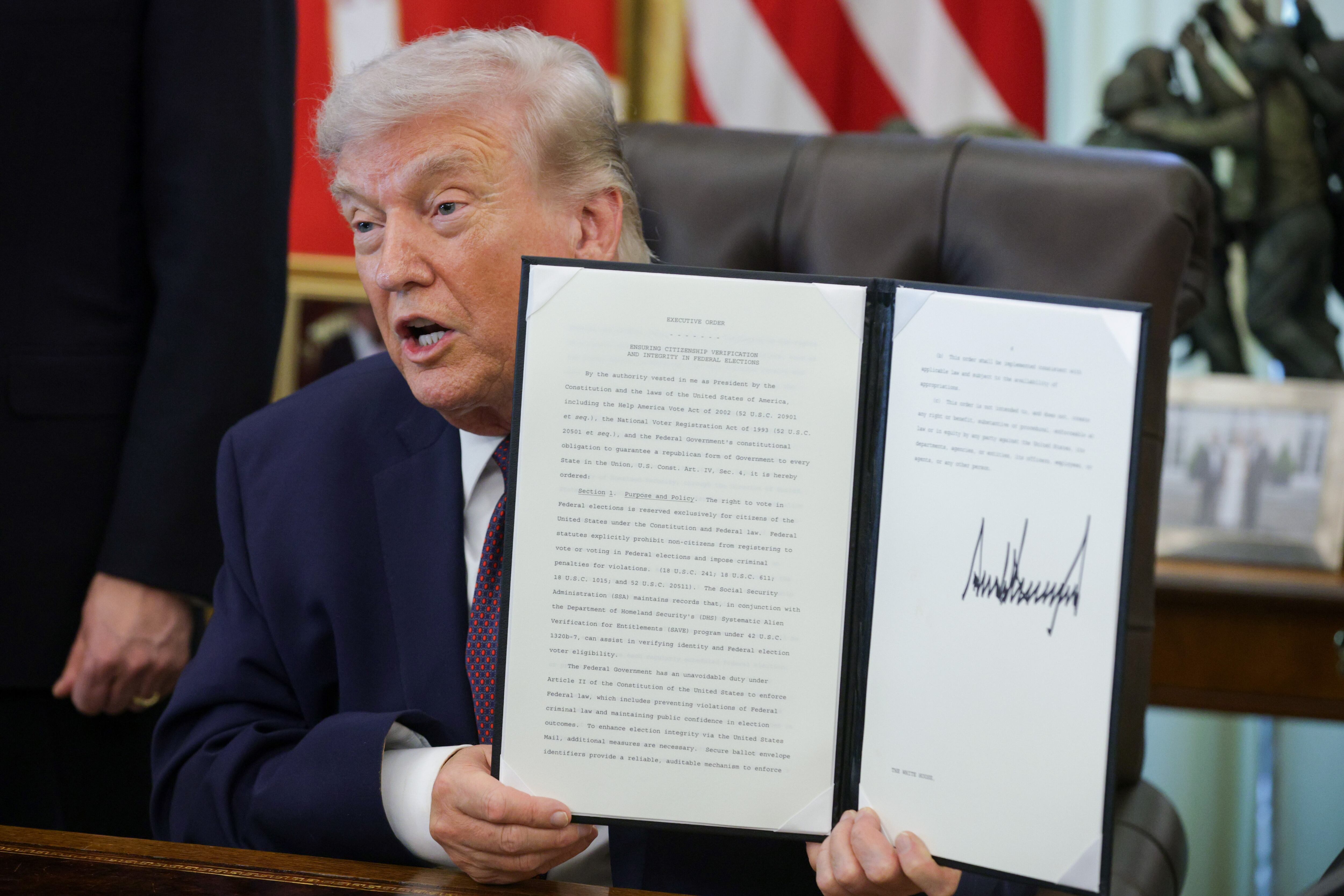 A man in a navy suit jacket holds a document at a desk.