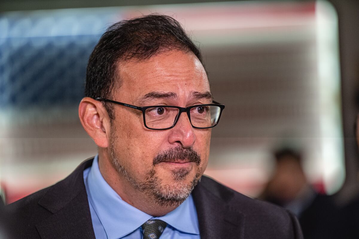 A man with a goatee and glasses looks away, with an American flag behind him