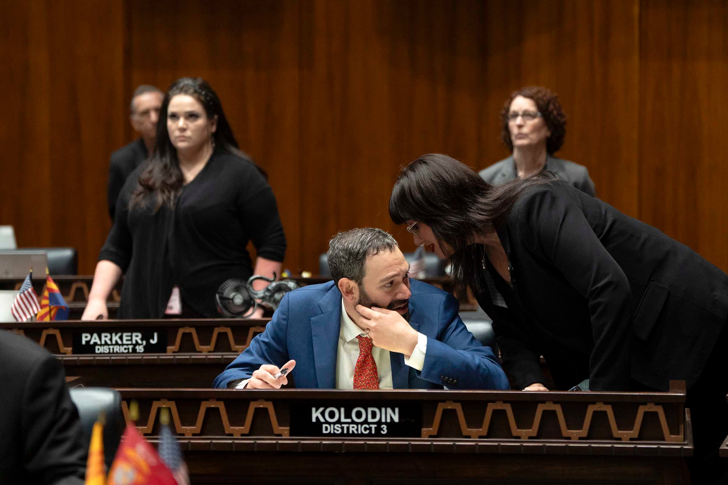 A photograph of two white people in suits talking to each other in a large wooden room.