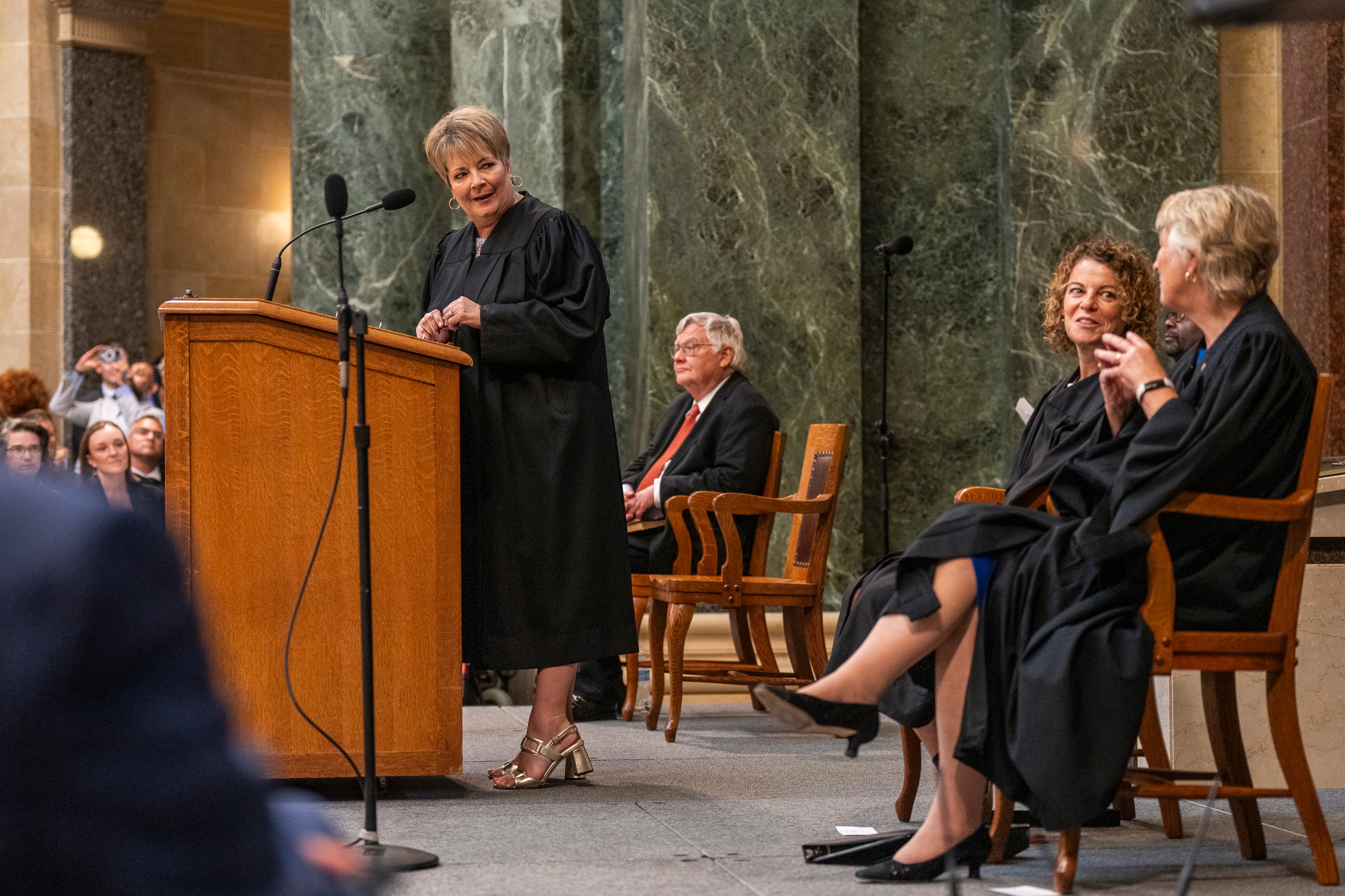A woman in a black robe stands behind a wooden podium next to three others sitting in black robes.