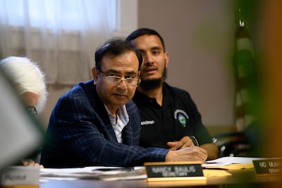 Three people sit side by side at a table in a conference room. The woman on the far left is partially covered.