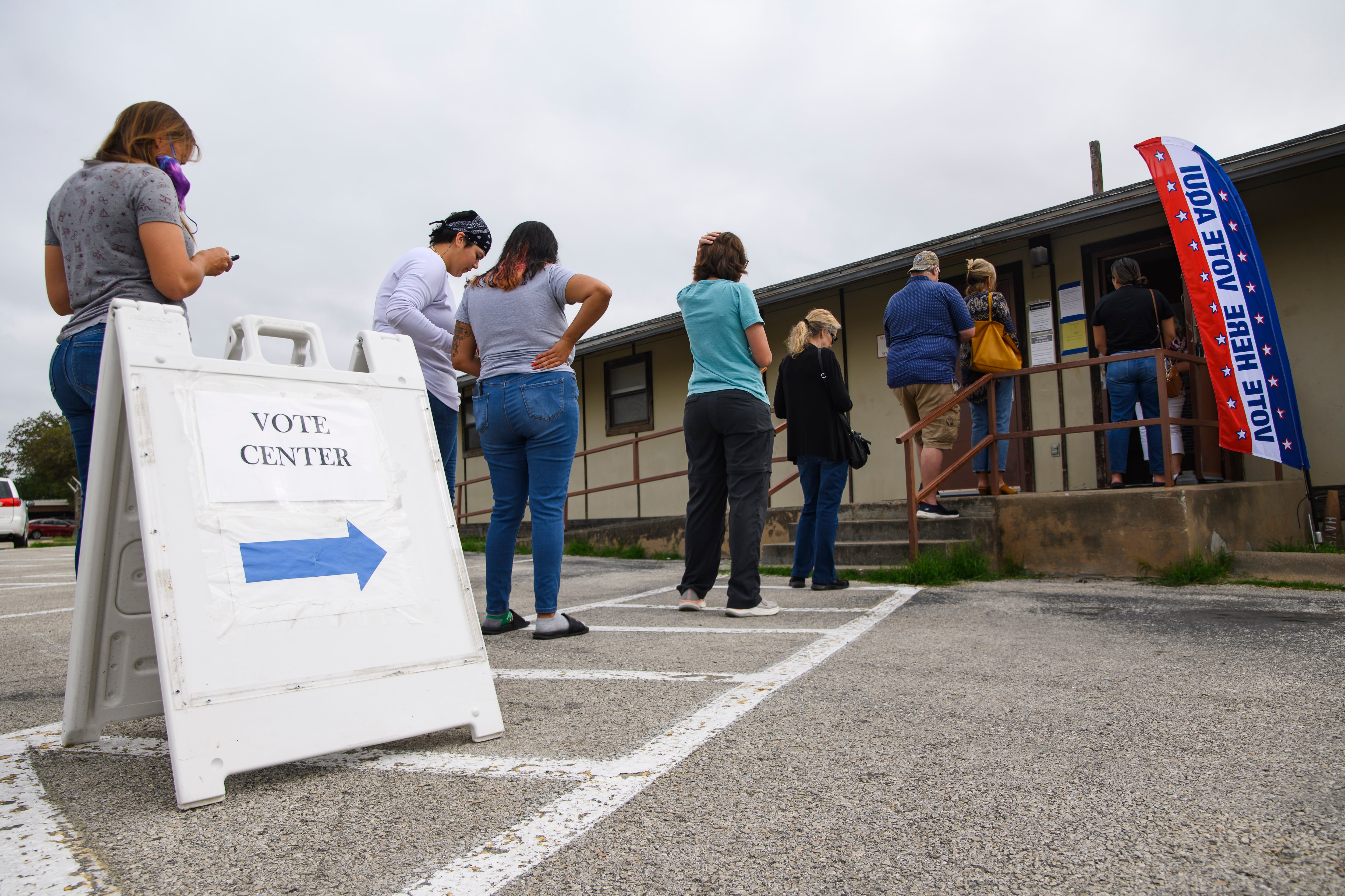 Several people stand in line in front of a building near a sign that says “vote center”