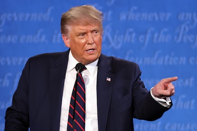 A man in a dark suit and a dark tie with red stripes stands and points in front of a blue background.